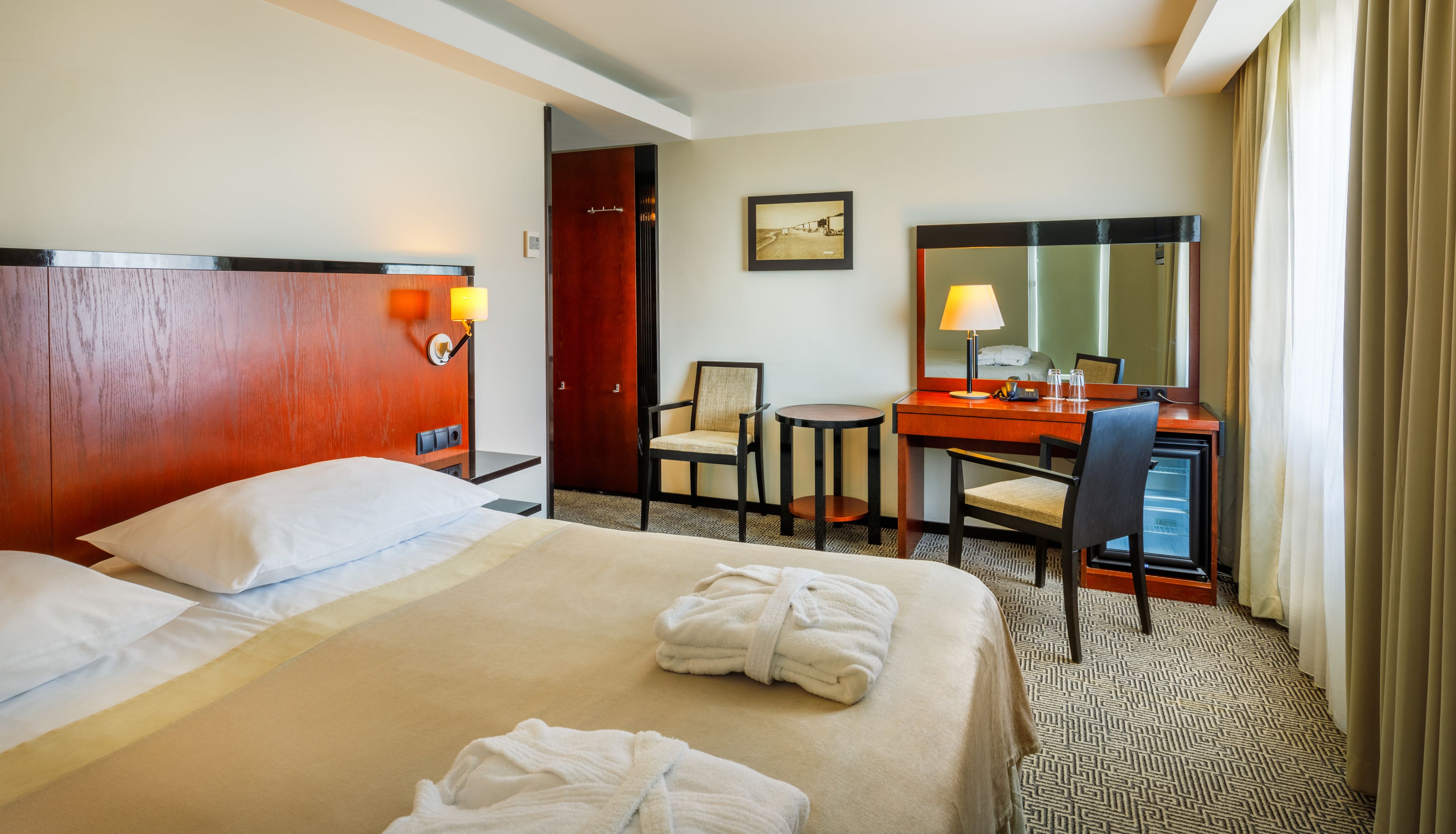 A hotel room with a bed, a desk, a chair, a small table, a mirror, and a picture on the wall.
