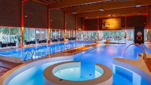 Une grande piscine intérieure avec un jacuzzi au centre, entourée de chaises longues et de tables.