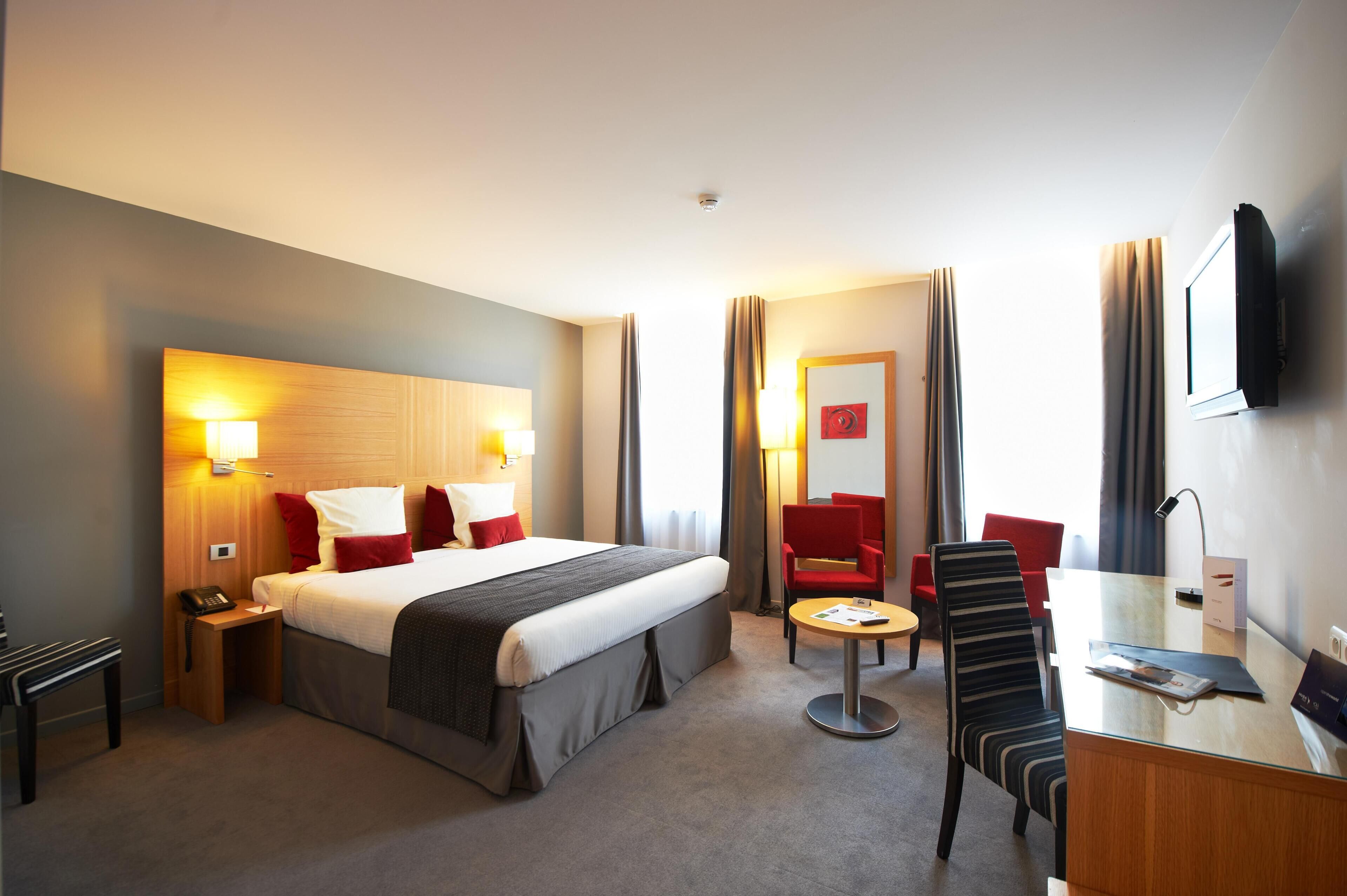 A hotel room with a bed, bedside tables, a desk with a chair, a TV, and a red chair.