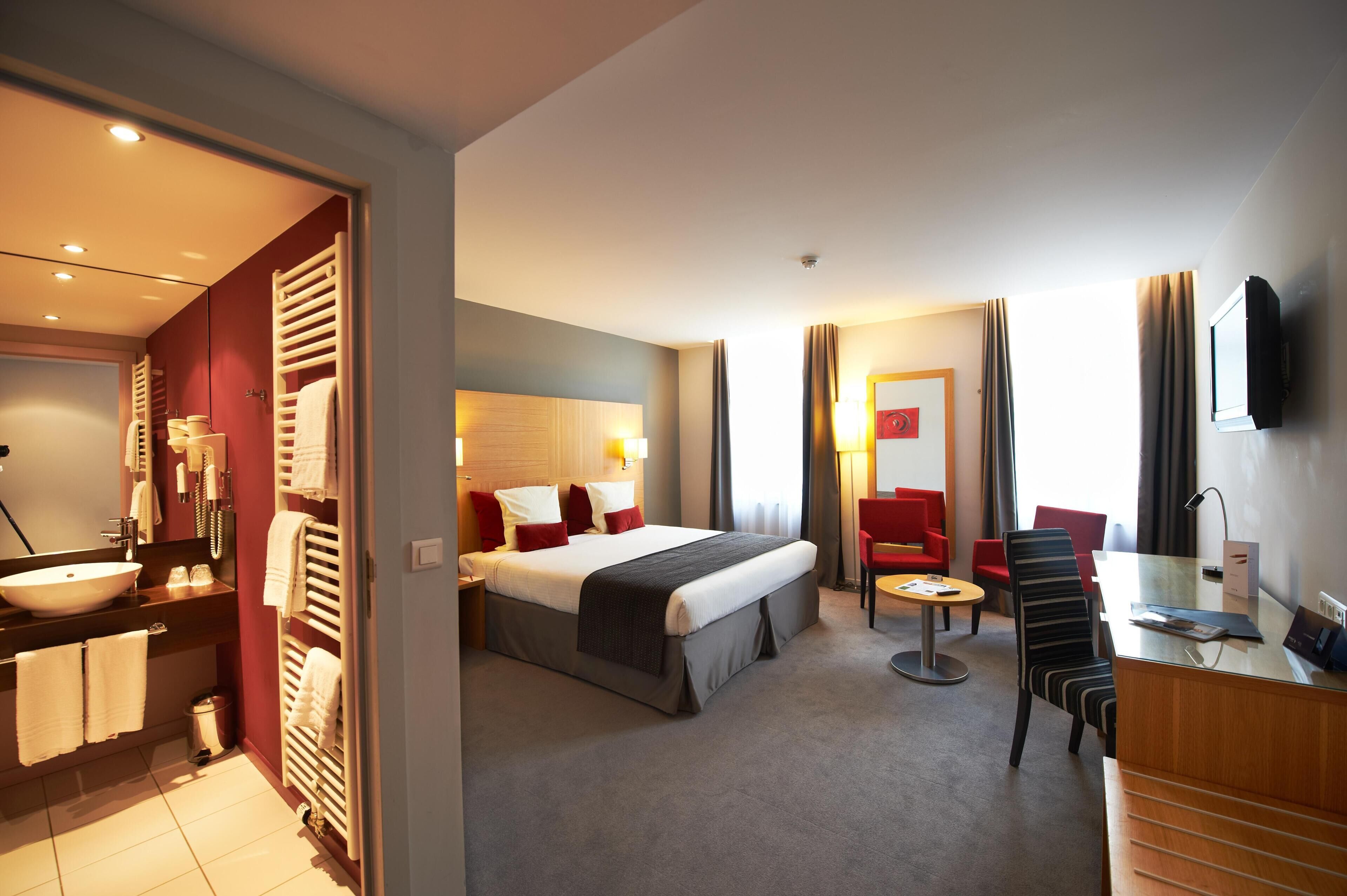 A hotel room with a bed, bedside table, desk, chair, and a bathroom with a sink and towel rack.