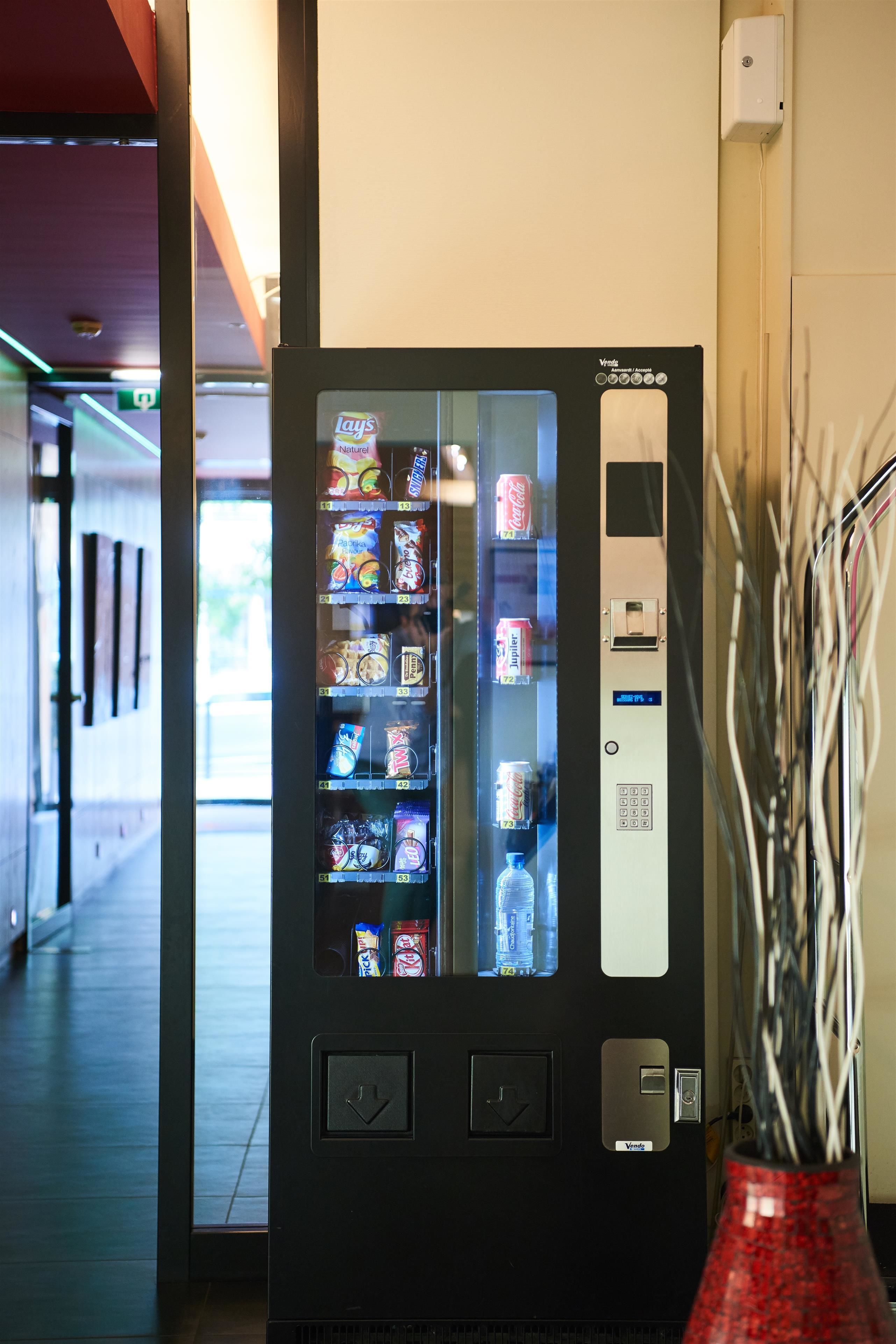 A vending machine with various snacks and drinks, located in a hallway with a red vase and some wires on the wall.