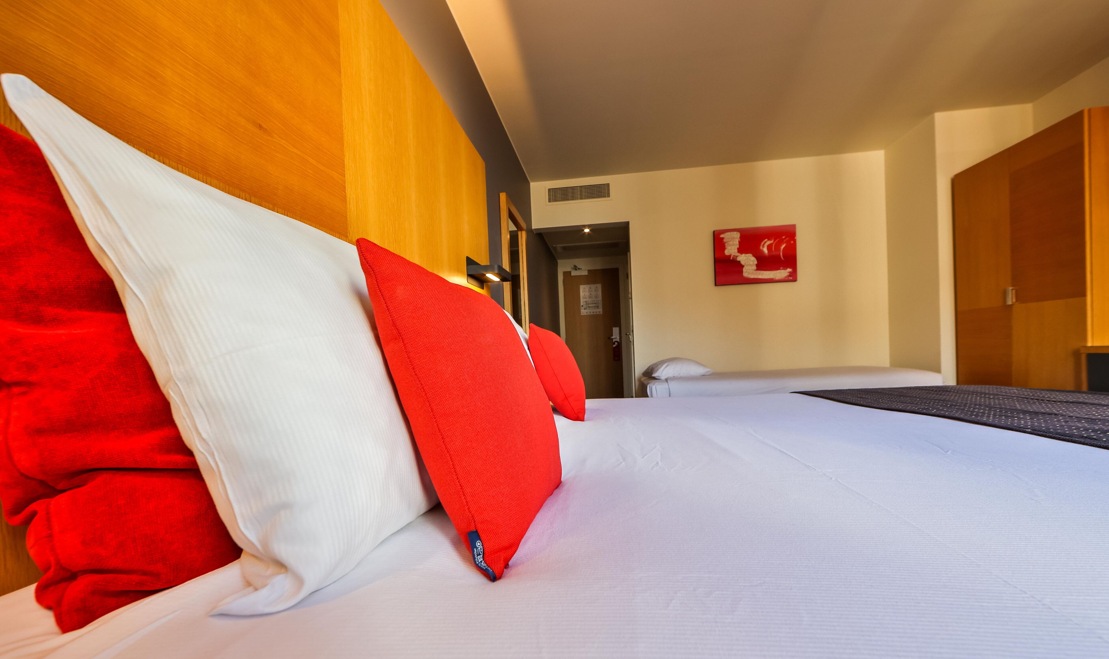 A hotel room with a bed, a wooden headboard, and a red and white pillow.