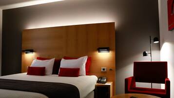 Une chambre d’hôtel avec un grand lit, des oreillers rouges et blancs, une tête de lit en bois, des lampes de chevet, une table de chevet avec un téléphone et une chaise rouge.