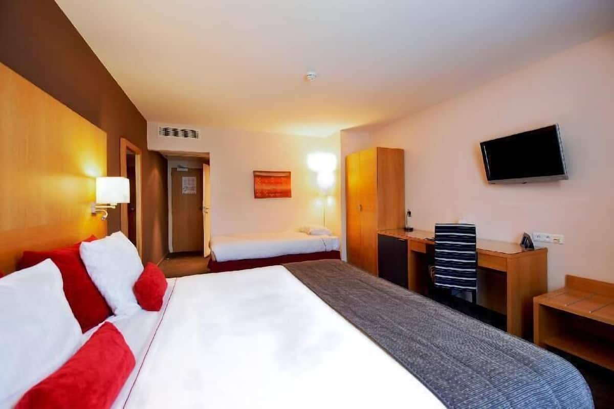 A hotel room with a large bed, a smaller bed, a desk, a chair, a TV, and a wall-mounted air conditioning unit.