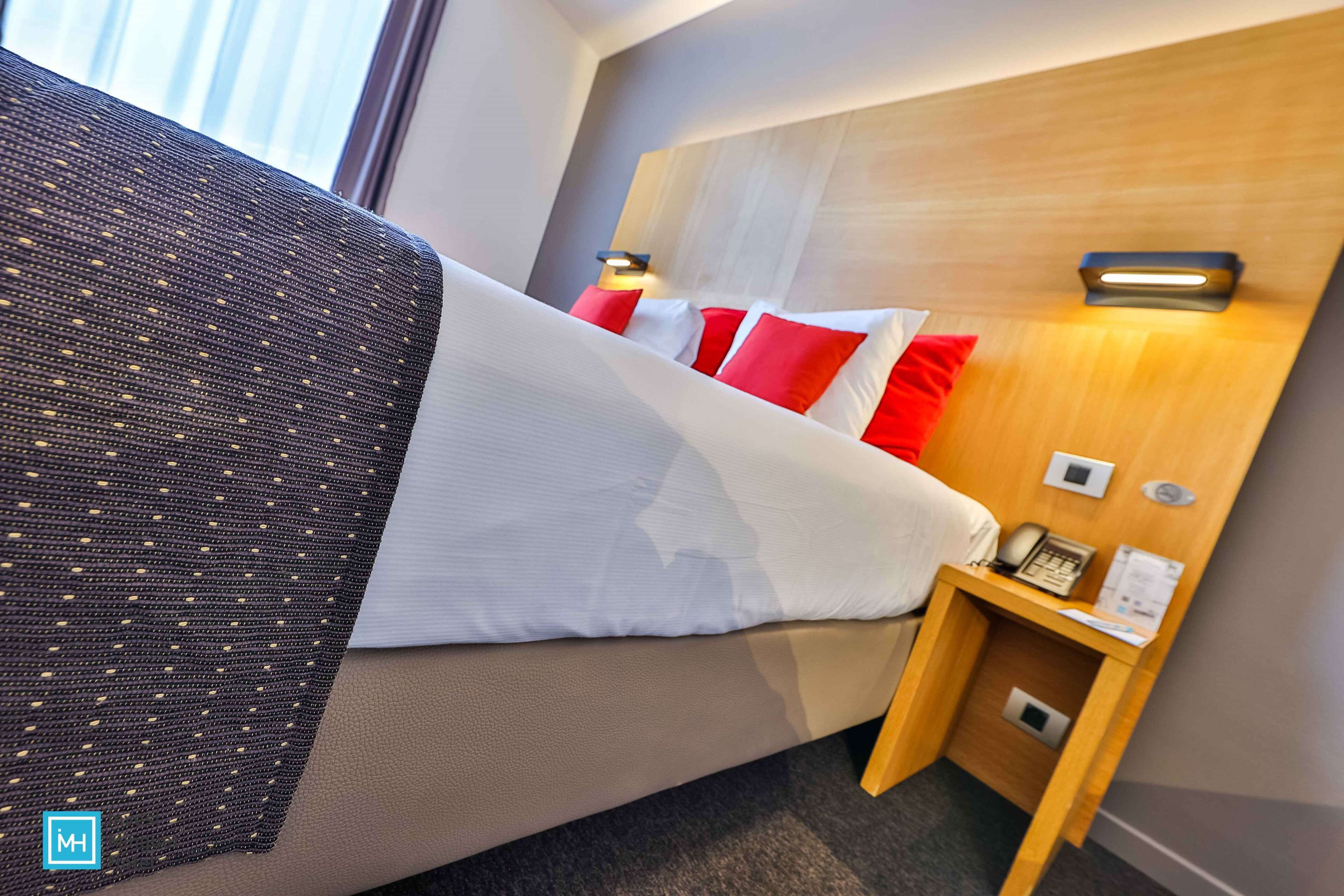 A hotel room with a bed, a nightstand with a phone and a lamp, and a headboard with built-in lighting.