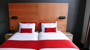 Een hotelkamer met een houten hoofdeinde, twee bedden met wit en rode beddengoed, nachtkastjes met zwarte lampen, en een telefoon op de linker tafel.