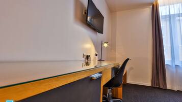 A hotel room with a glass desk, a black chair, a mounted flat-screen TV, a bedside table with a lamp, and a window with curtains.