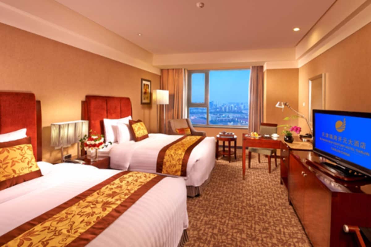 A hotel room with two beds, a desk, a television, and a view of the city.