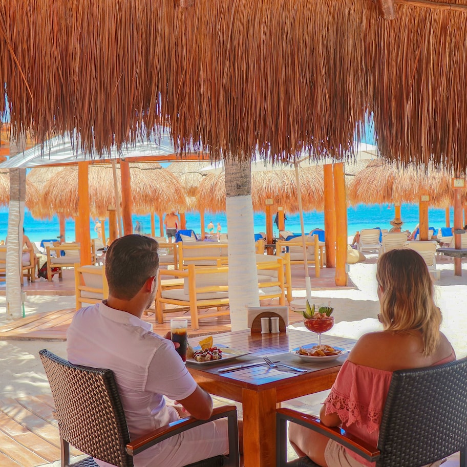 A beachside restaurant with thatched roofs, wooden tables, and chairs, overlooking the ocean.