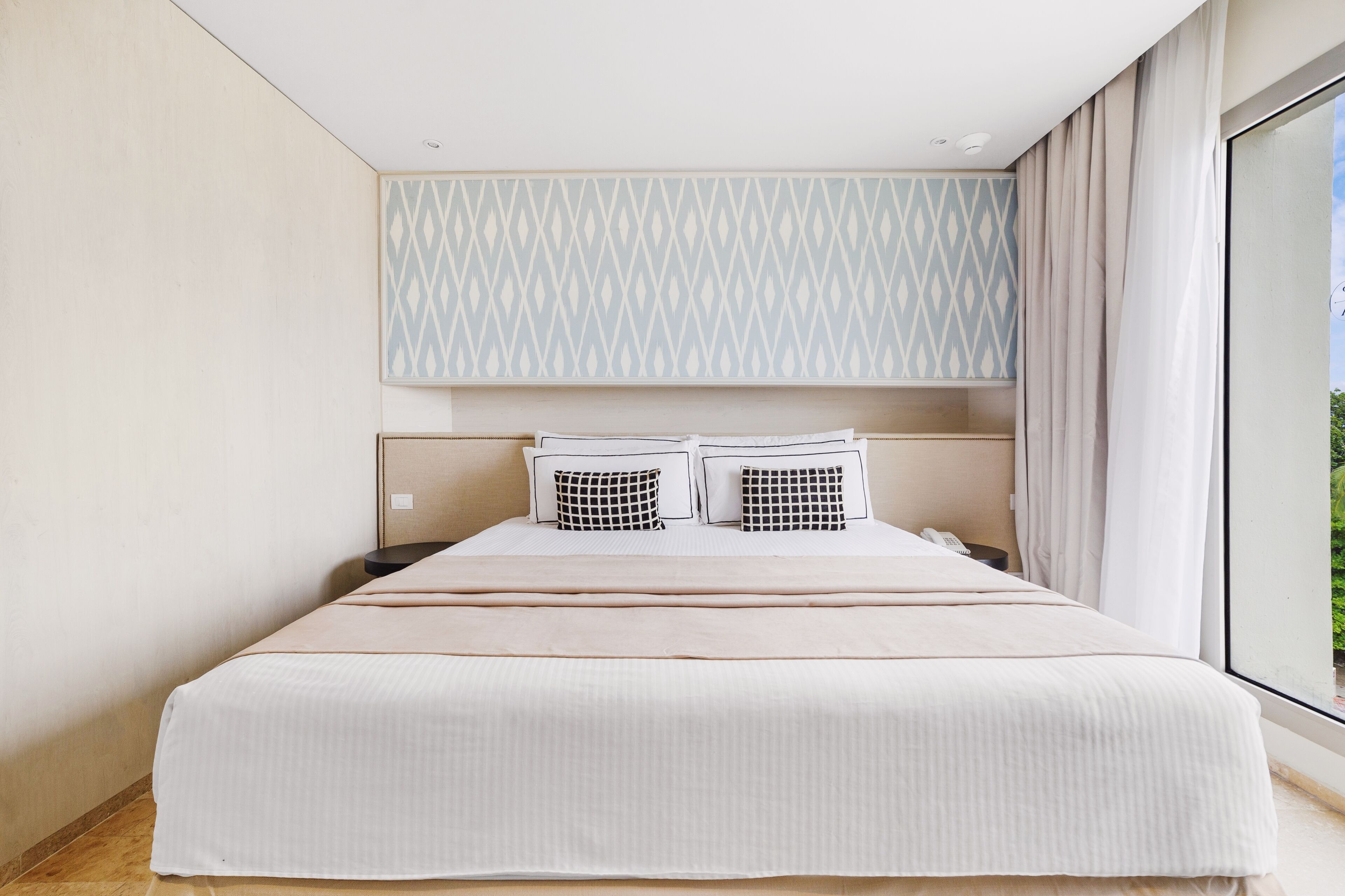 Quarto de hotel moderno com uma cama grande, cabeceira estampada e vista para o exterior.