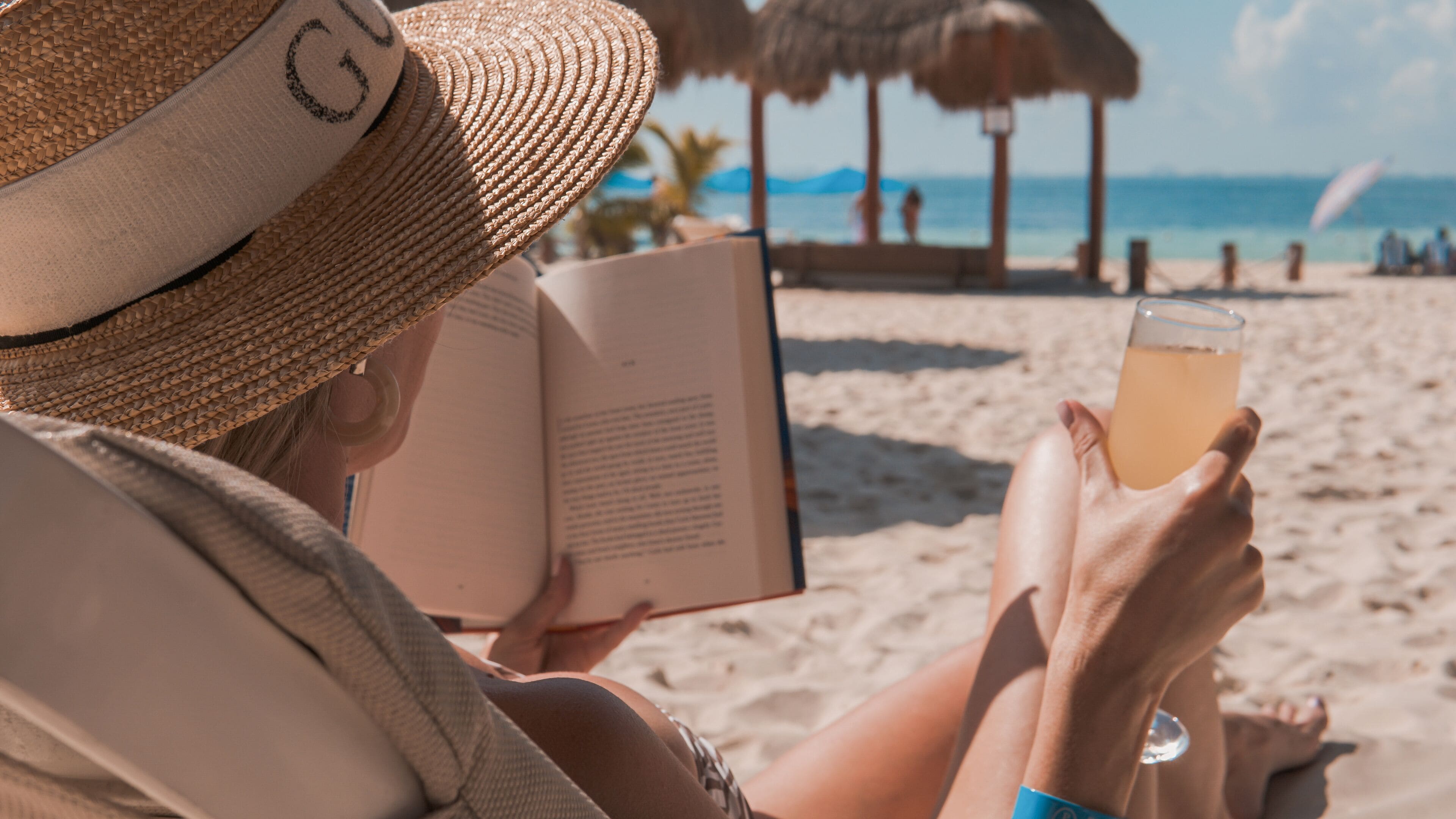 A person reading a book on a beach with a drink in hand.