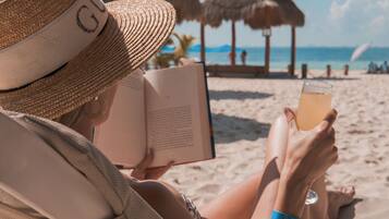 A person reading a book on a beach with a drink in hand.