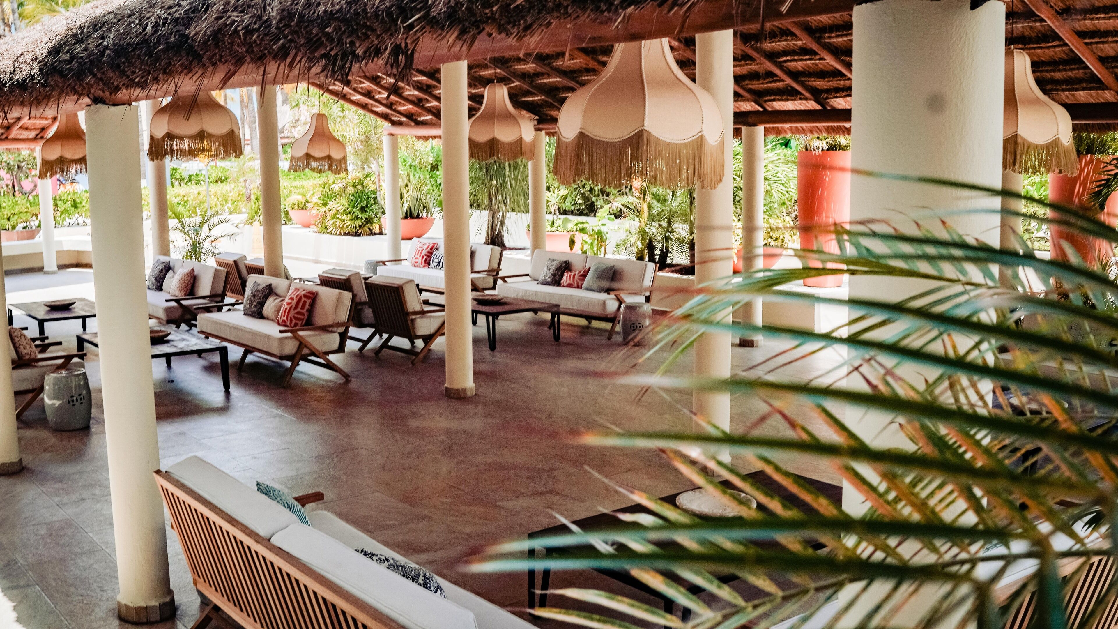 A covered outdoor seating area with multiple sofas and tables, surrounded by potted plants and a thatched roof.