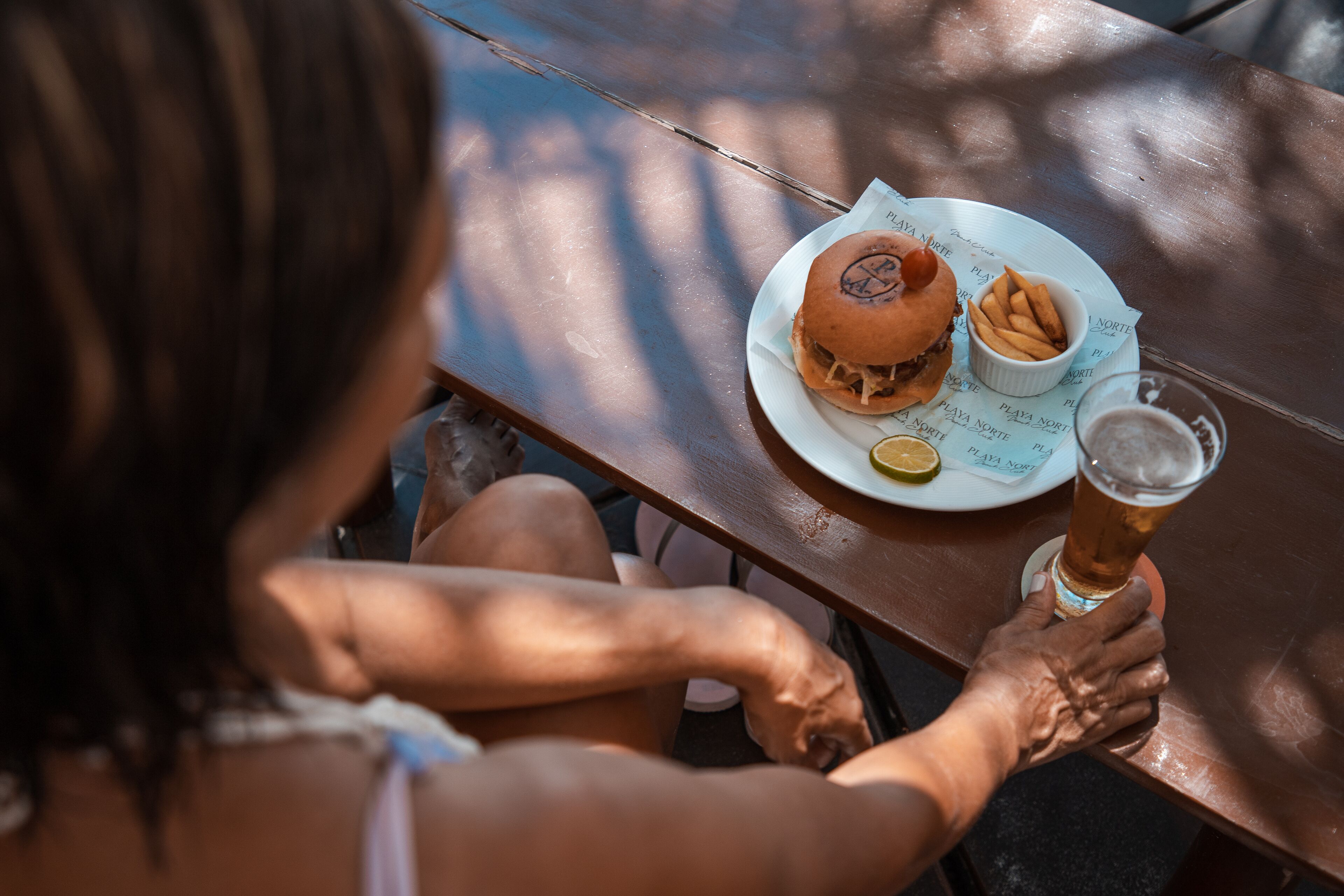 Une personne est assise à une table en bois, devant une assiette de nourriture et un verre de bière.