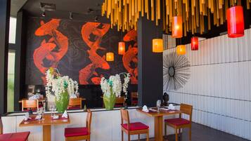 Interior de restaurante con mesas y sillas de madera, barra de sushi y un mural de peces rojos.