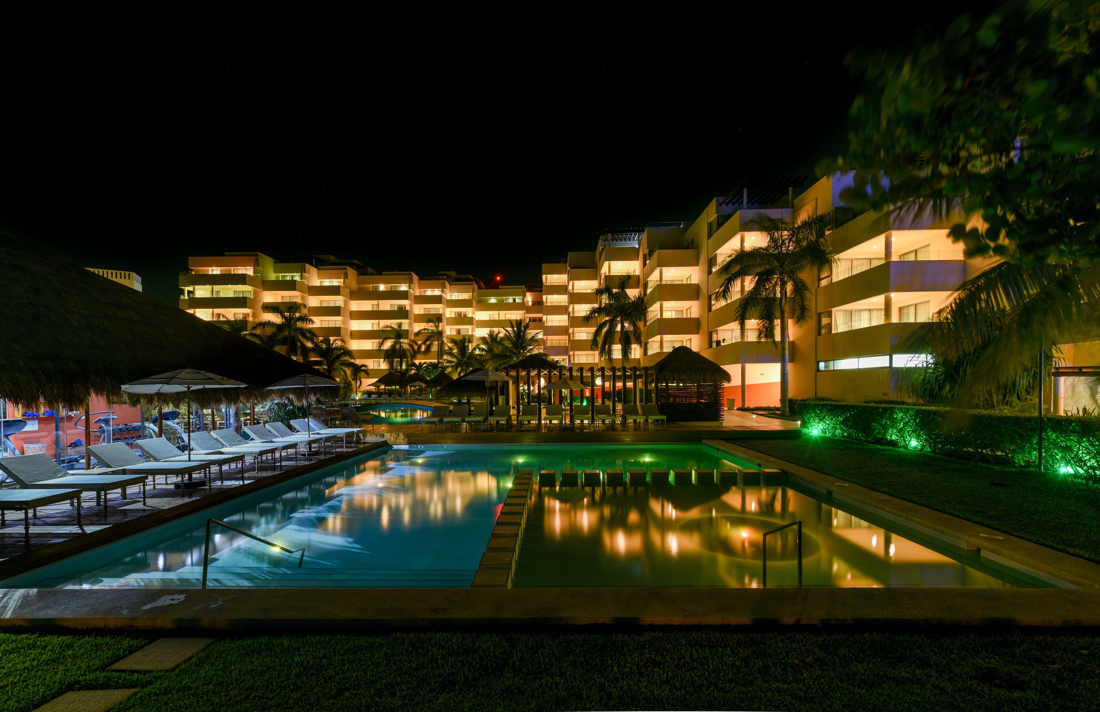 A nighttime resort pool area with lounge chairs and a reflection pool, surrounded by hotel buildings and palm trees.