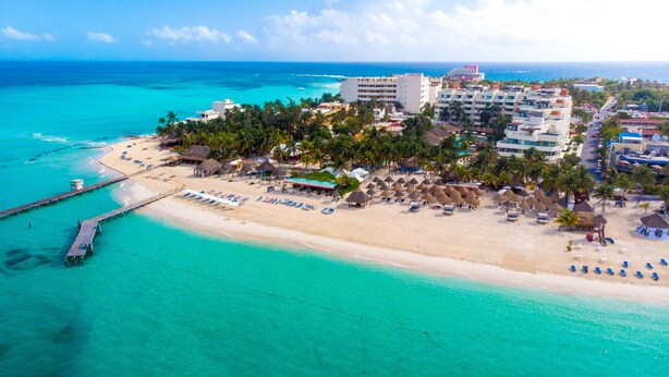 Vista aérea de un complejo turístico en la playa con muelle, sombrillas y un hotel de varios pisos.