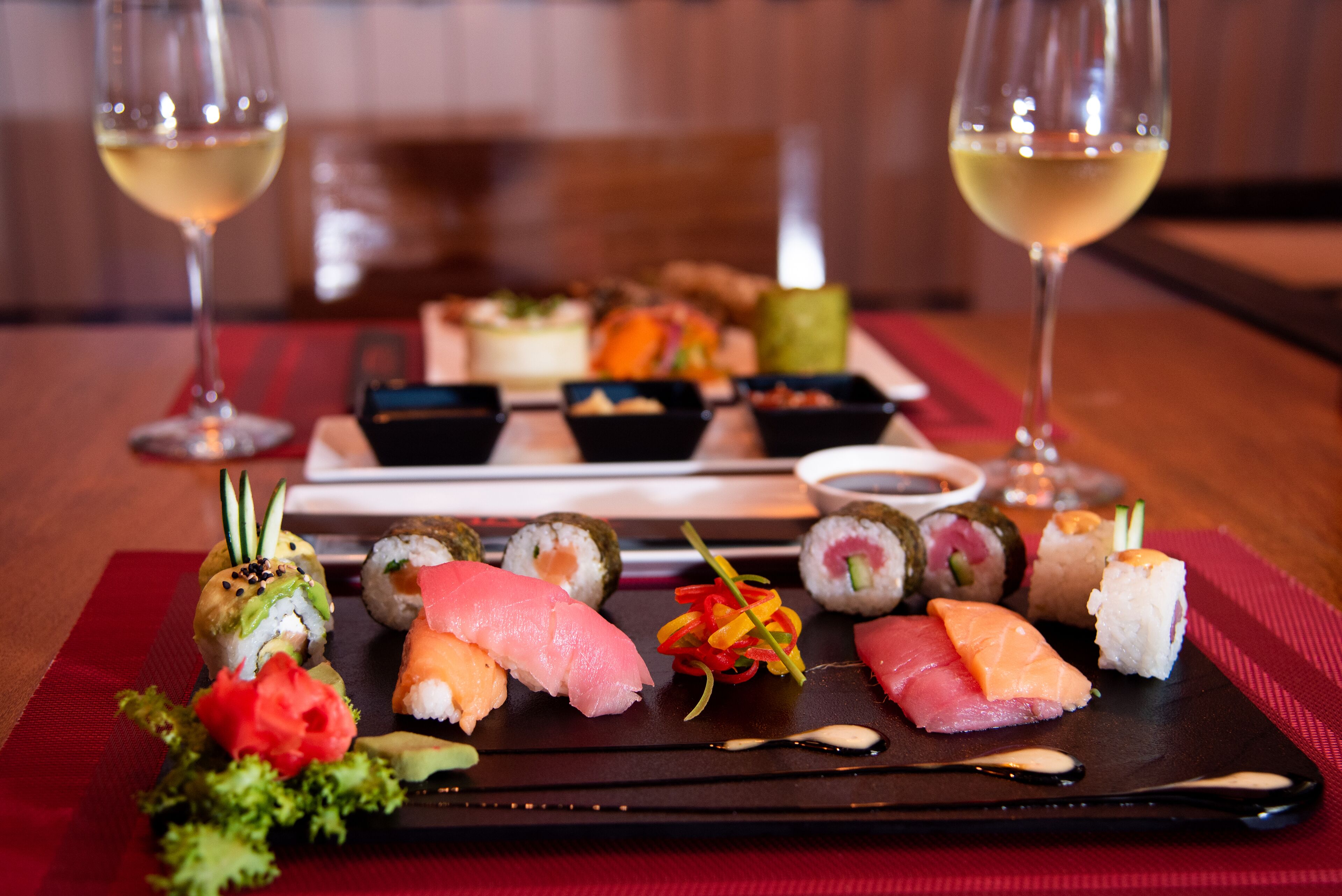 A variety of sushi on a black tray with a red placemat, accompanied by two glasses of white wine on a wooden table.