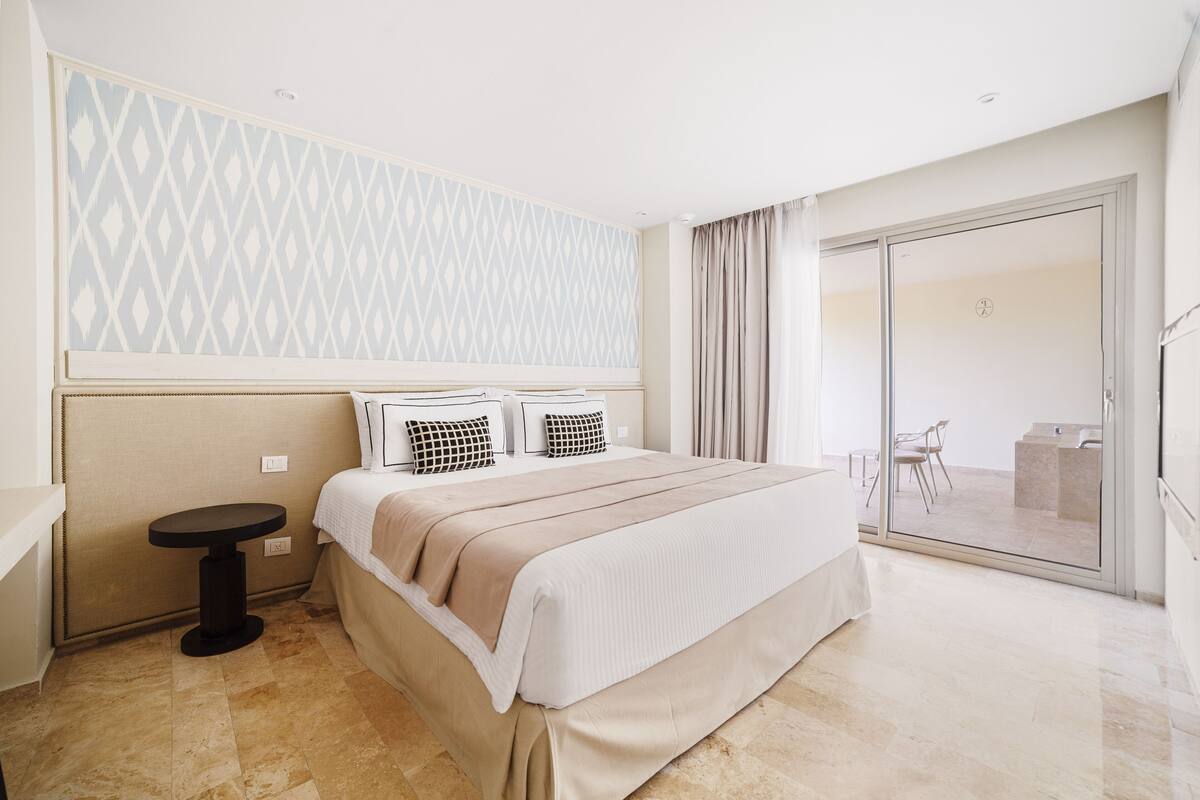 Ein Hotelzimmer mit einem Bett, einem kleinen runden Tisch, einer Wand mit Muster und einer Schiebeglastür zu einem Balkon.