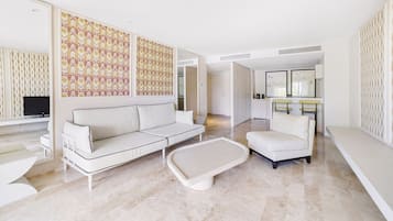 A modern living room with a white sofa, a white armchair, and a coffee table. There is a kitchen area in the background with a bar and stools.