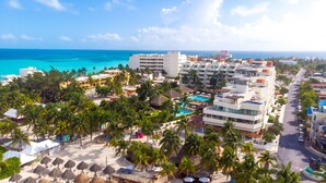 Un resort con varias edificaciones, un área de piscina y una playa con sombrillas y palmeras.