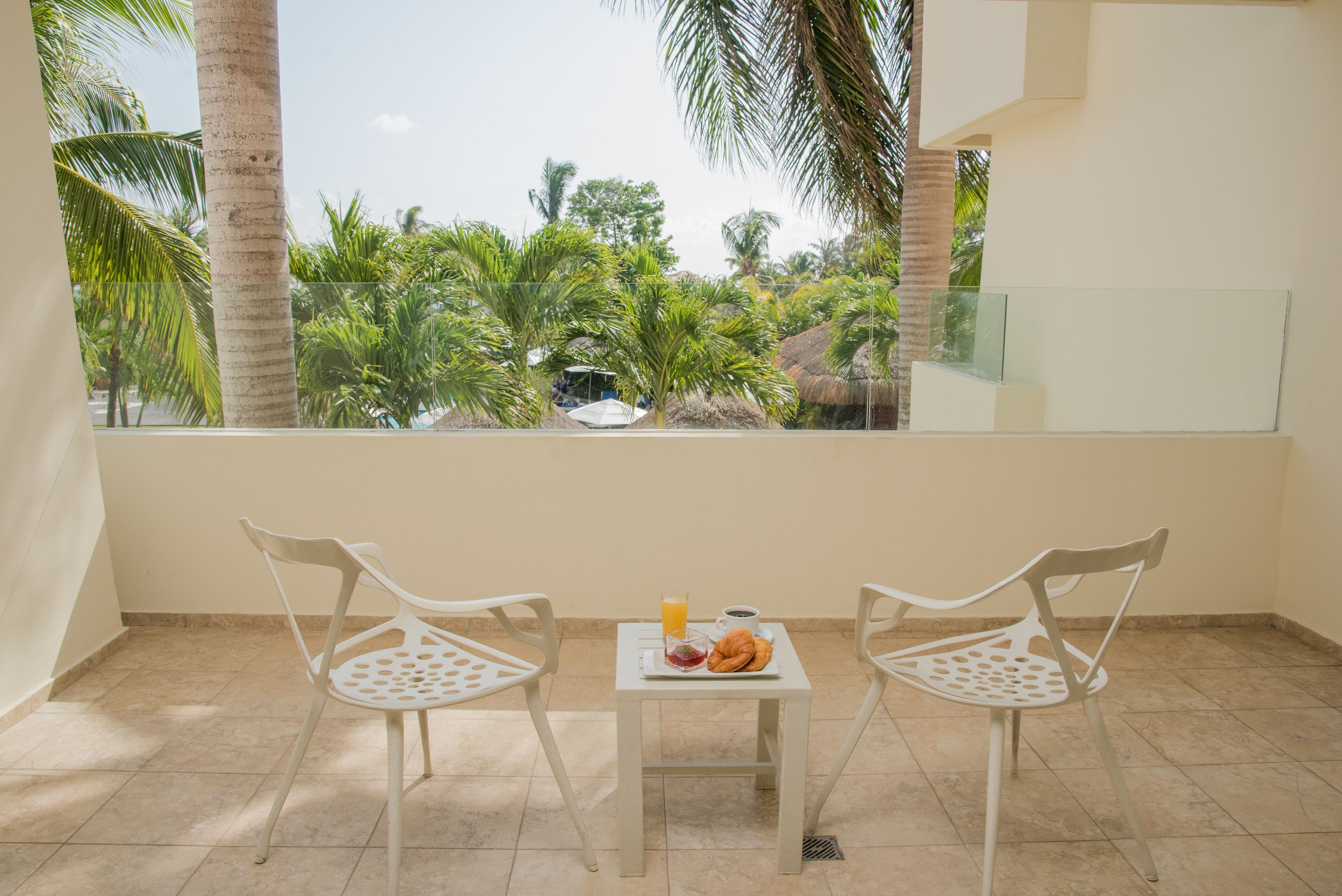 A balcony with two white chairs, a small table with breakfast items, and a view of palm trees and a thatched-roof structure.