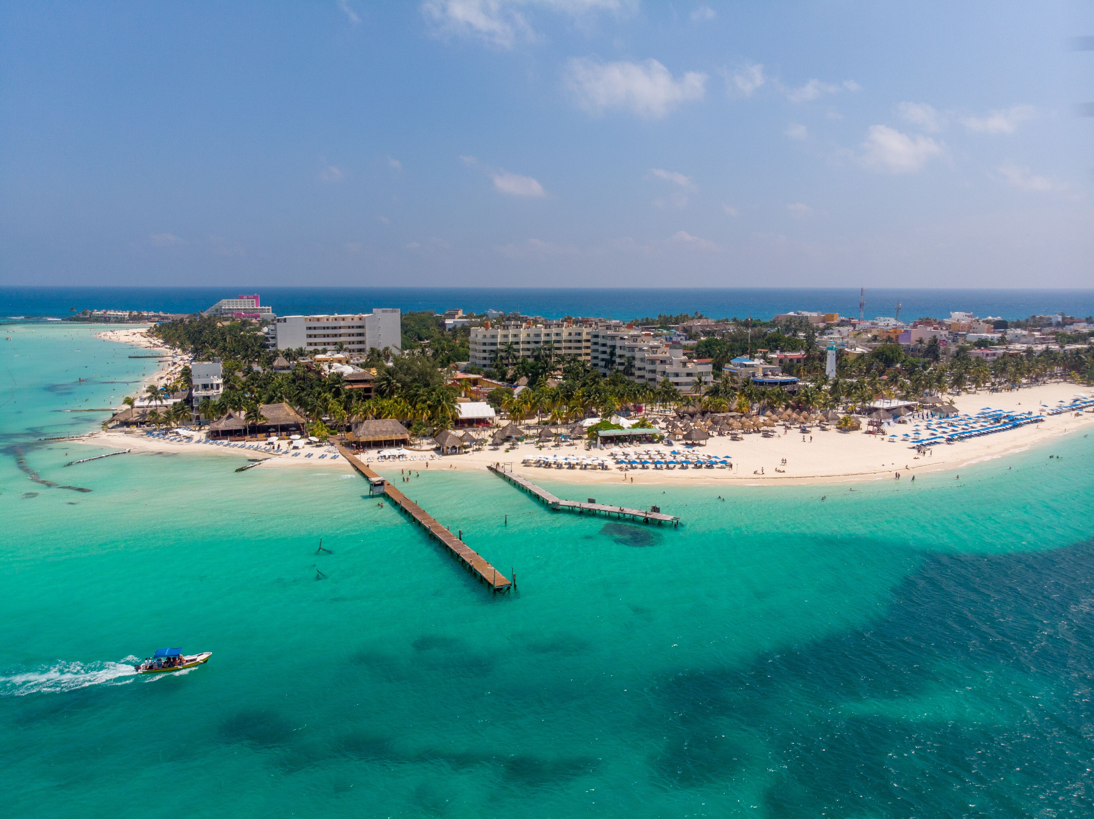 An aerial view of a coastal resort with a pier extending into turquoise waters, surrounded by beach umbrellas and a variety of buildings.