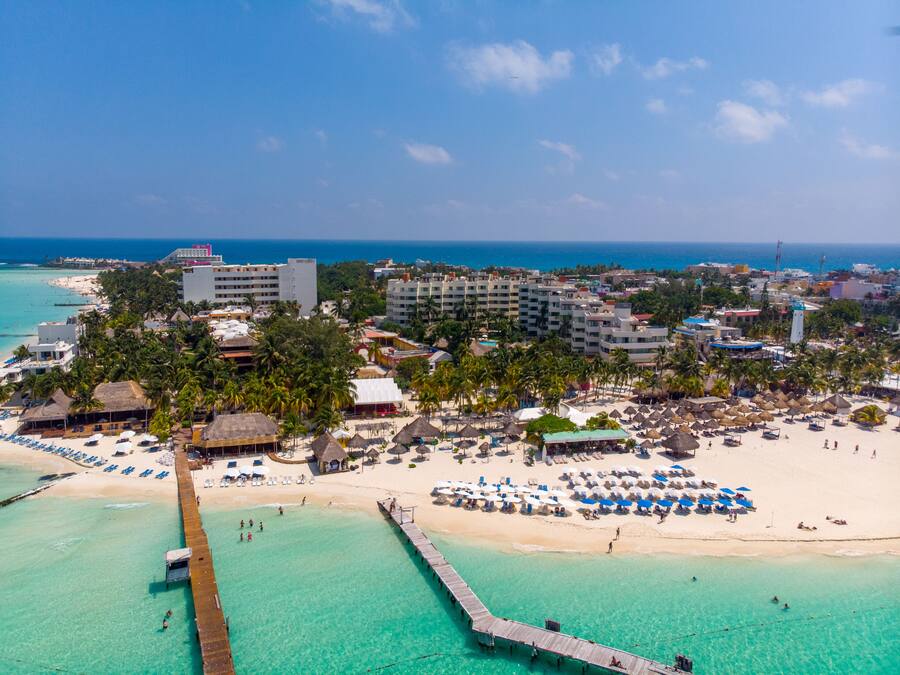 Un complejo turístico costero con un muelle que se adentra en aguas turquesas, villas frente a la playa y una playa arenosa con sombrillas.