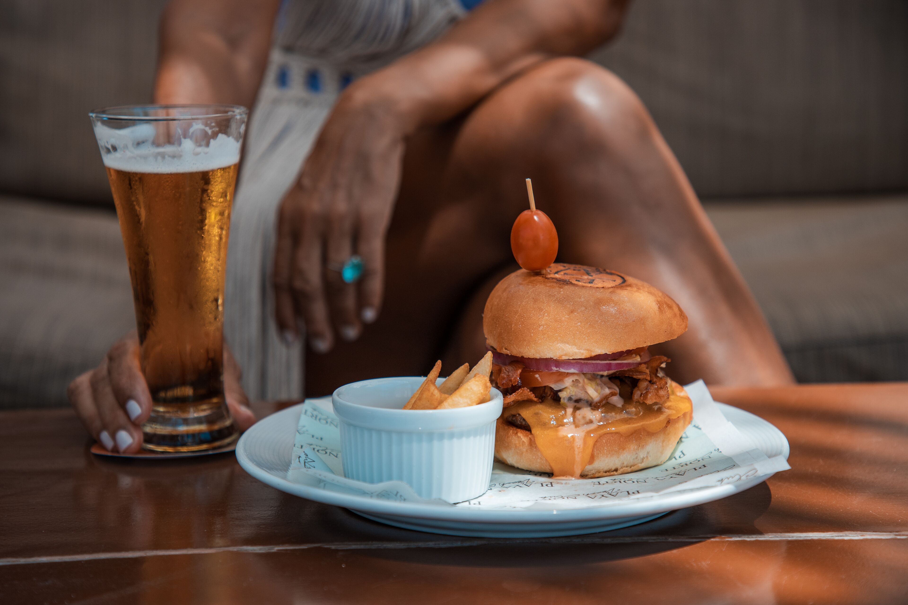 A plate with a burger and fries, a glass of beer, and a person sitting on a couch.