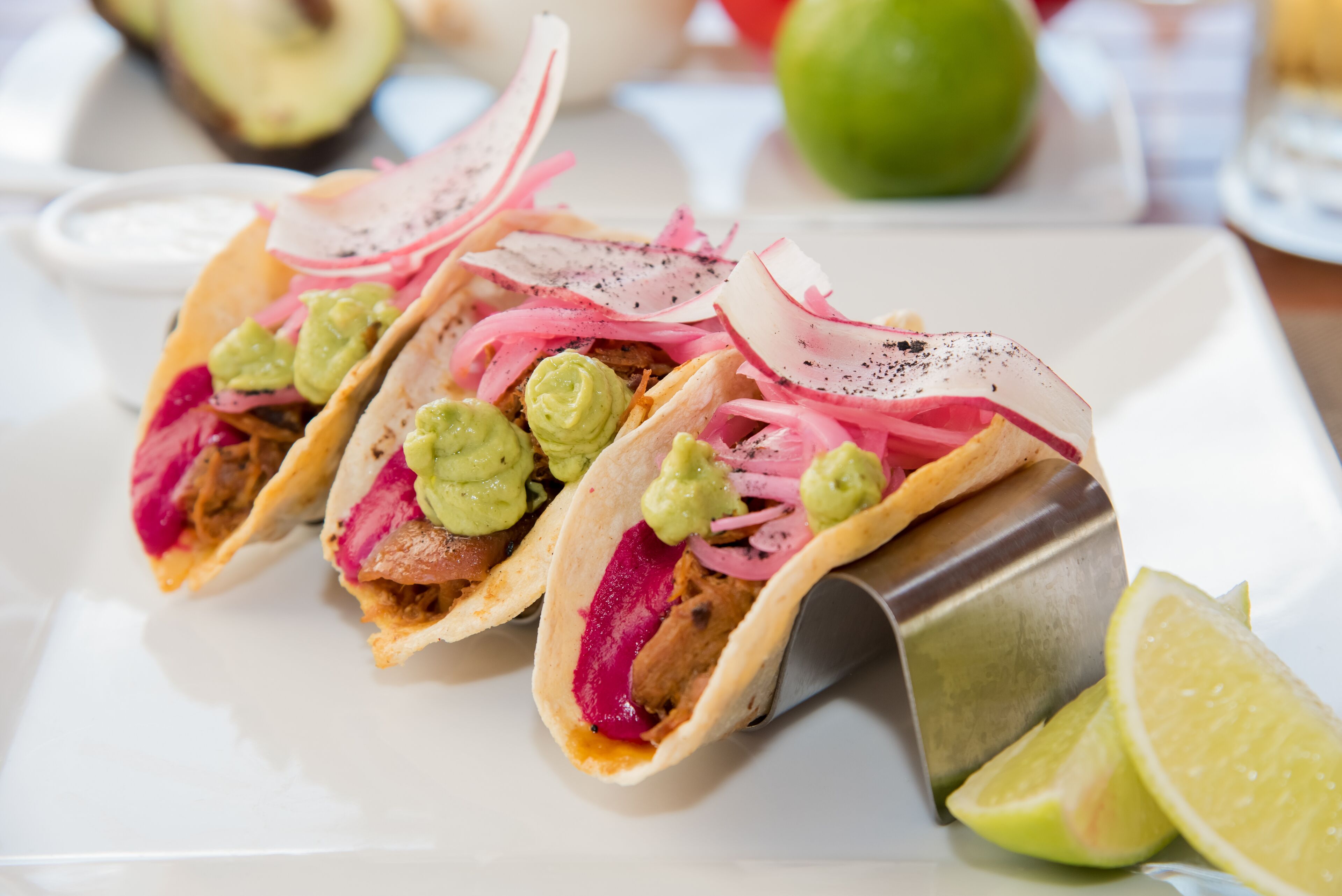 Tres tacos con distintos ingredientes sobre un plato blanco, acompañados de gajos de limón.