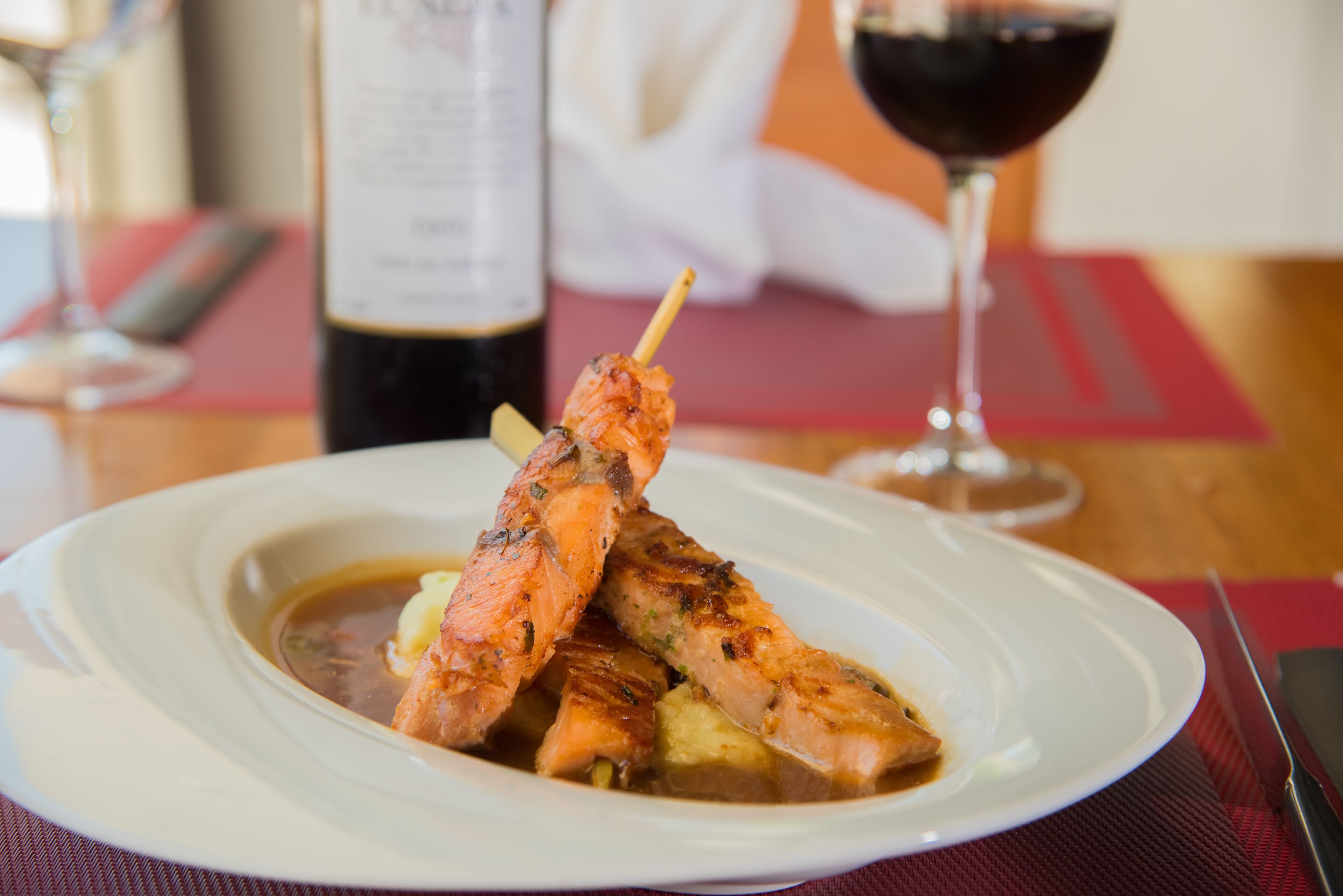 Un plato de salmón a la parrilla con patatas, una copa de vino tinto y una botella de vino sobre una mesa.