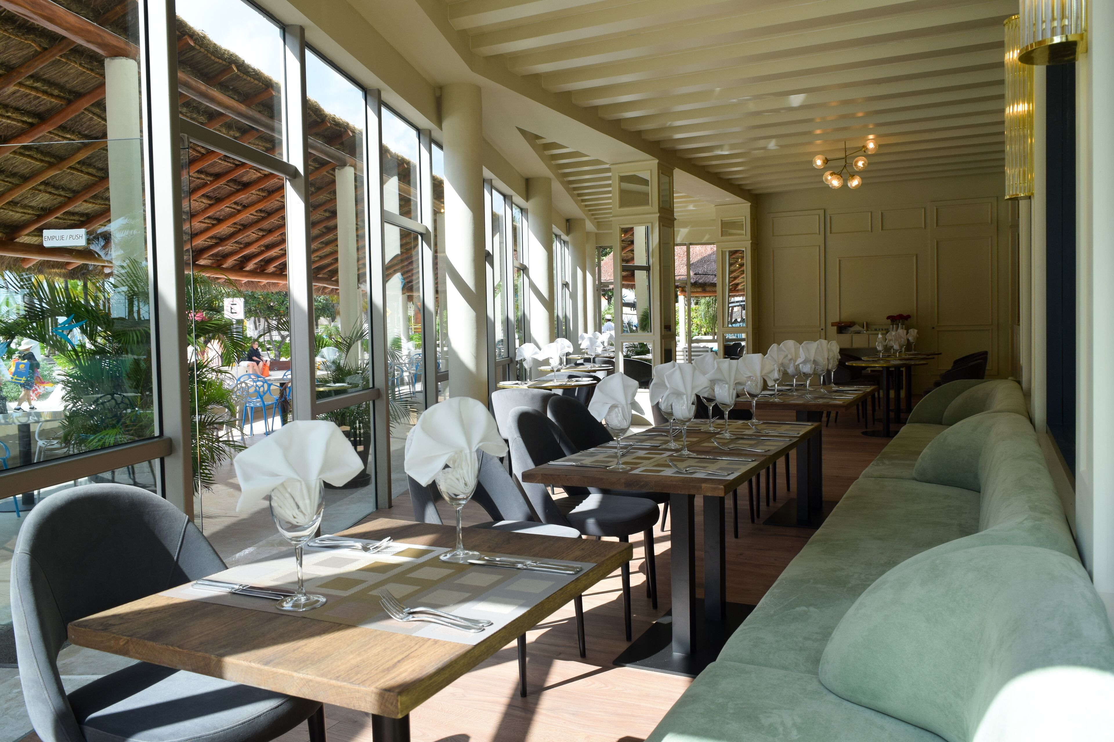 Un comedor interior con mesas dispuestas para dos, ventanas amplias y un largo banco a lo largo de una pared.
