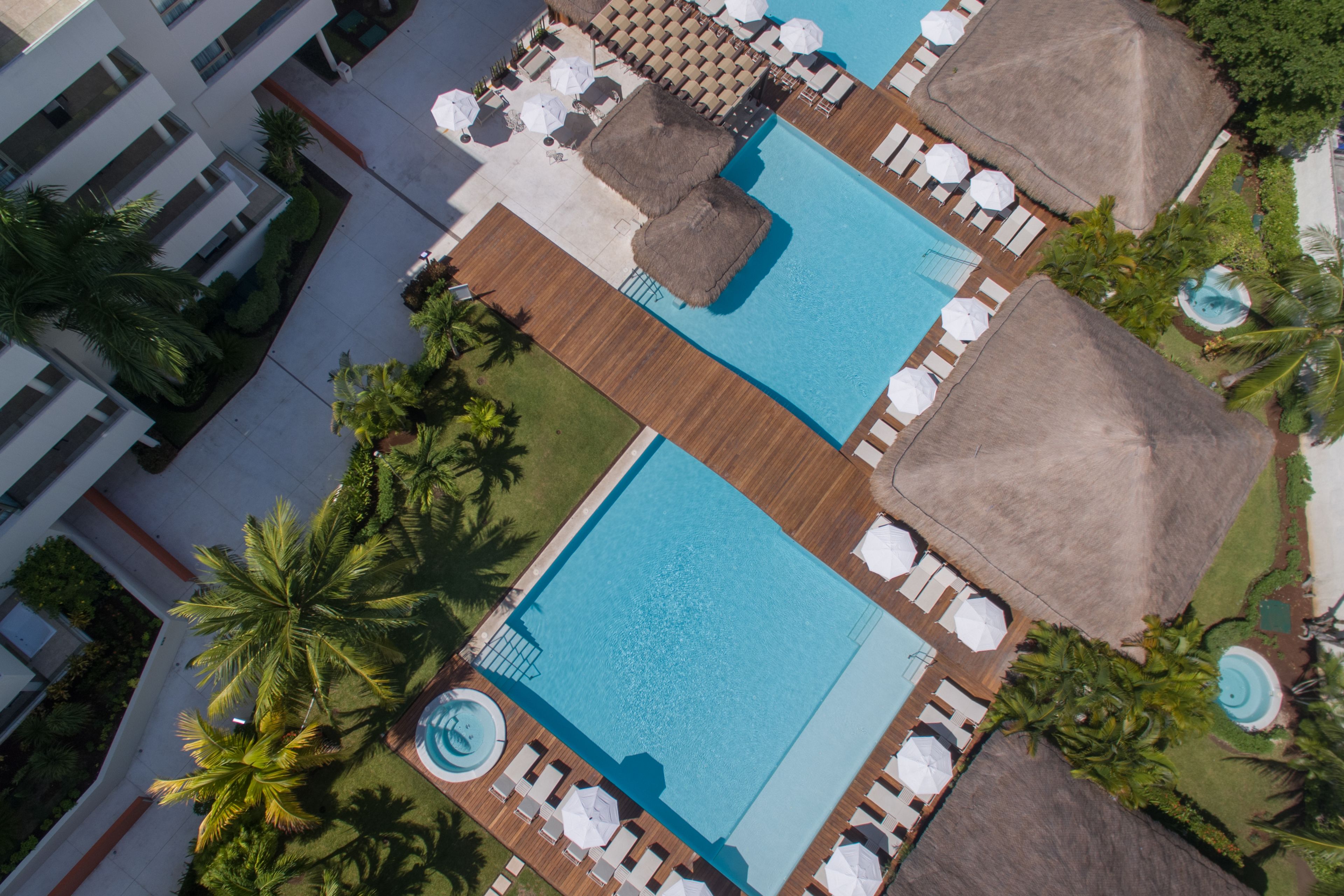 An aerial view of a resort with two large swimming pools, sun loungers, umbrellas, and palm trees.
