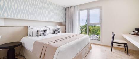 A modern bedroom with a large bed, a small table, a chair, and a view of greenery outside.