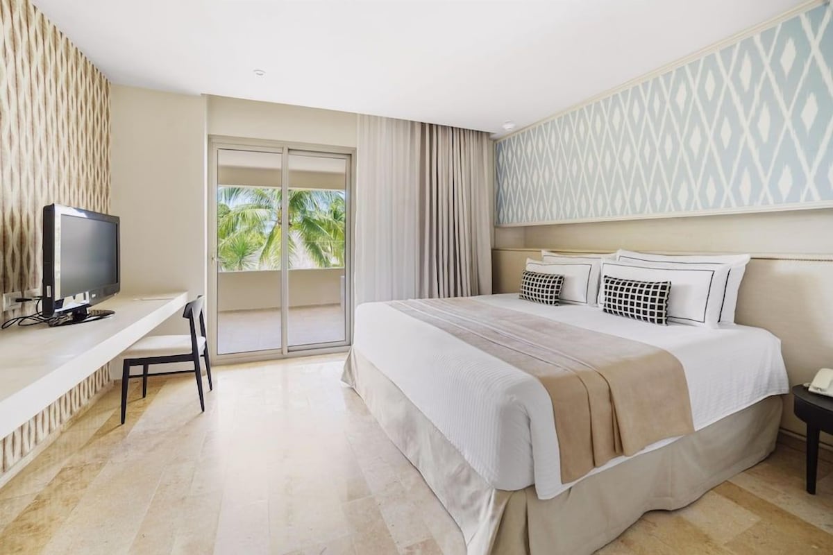 A hotel room with a bed, a desk with a television, a chair, and a view of palm trees through the window.