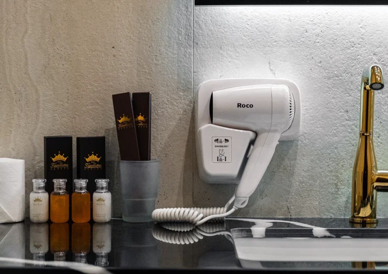Un baño con un secador de pelo montado en la pared, un grifo dorado y una fila de artículos de aseo, incluyendo botellas y una taza.