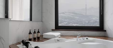 A modern bathroom with a large bathtub, a window with a snowy cityscape view, and toiletries on the side.