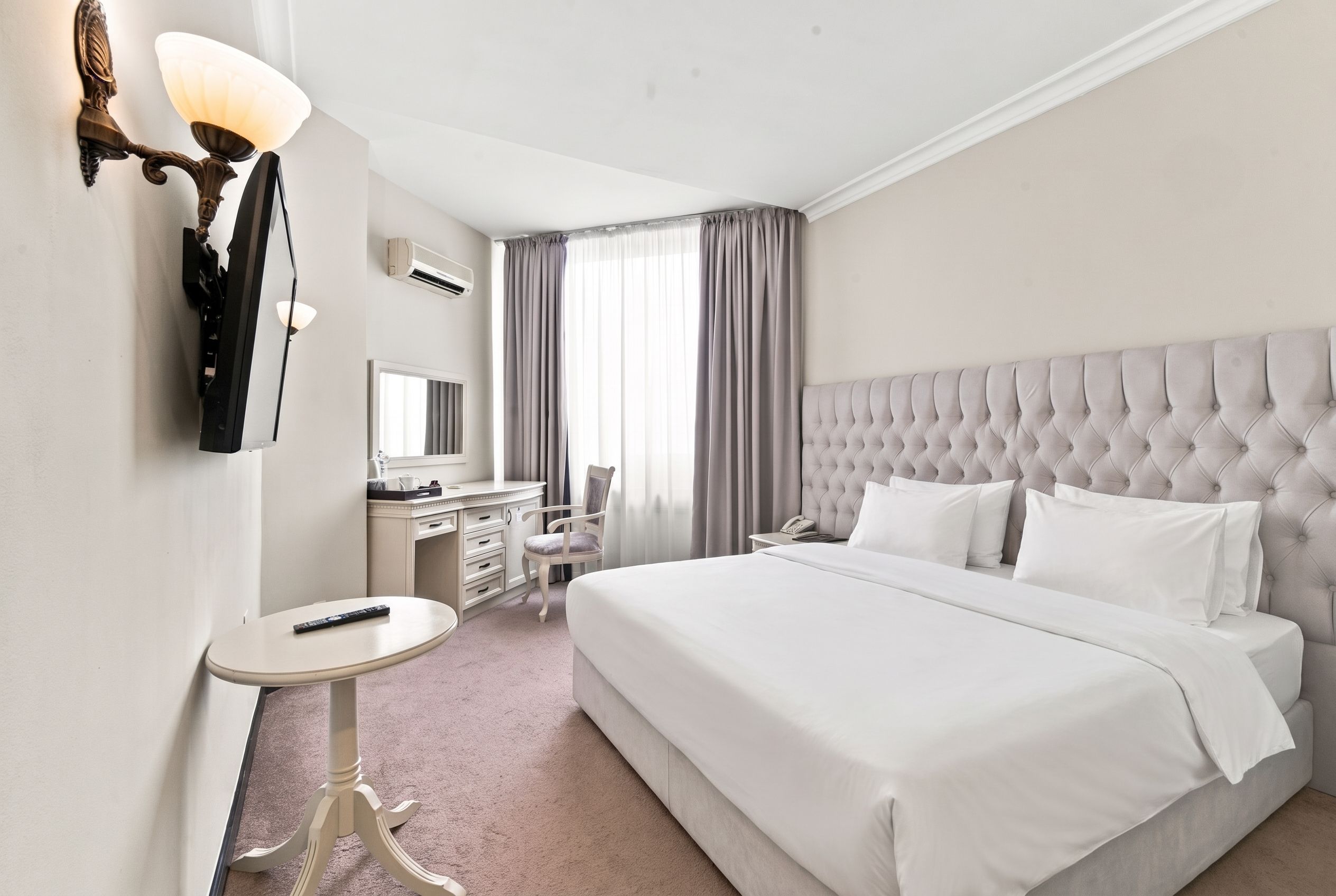 A hotel room with a large bed, a desk with a chair, a small round table, and a wall-mounted lamp.