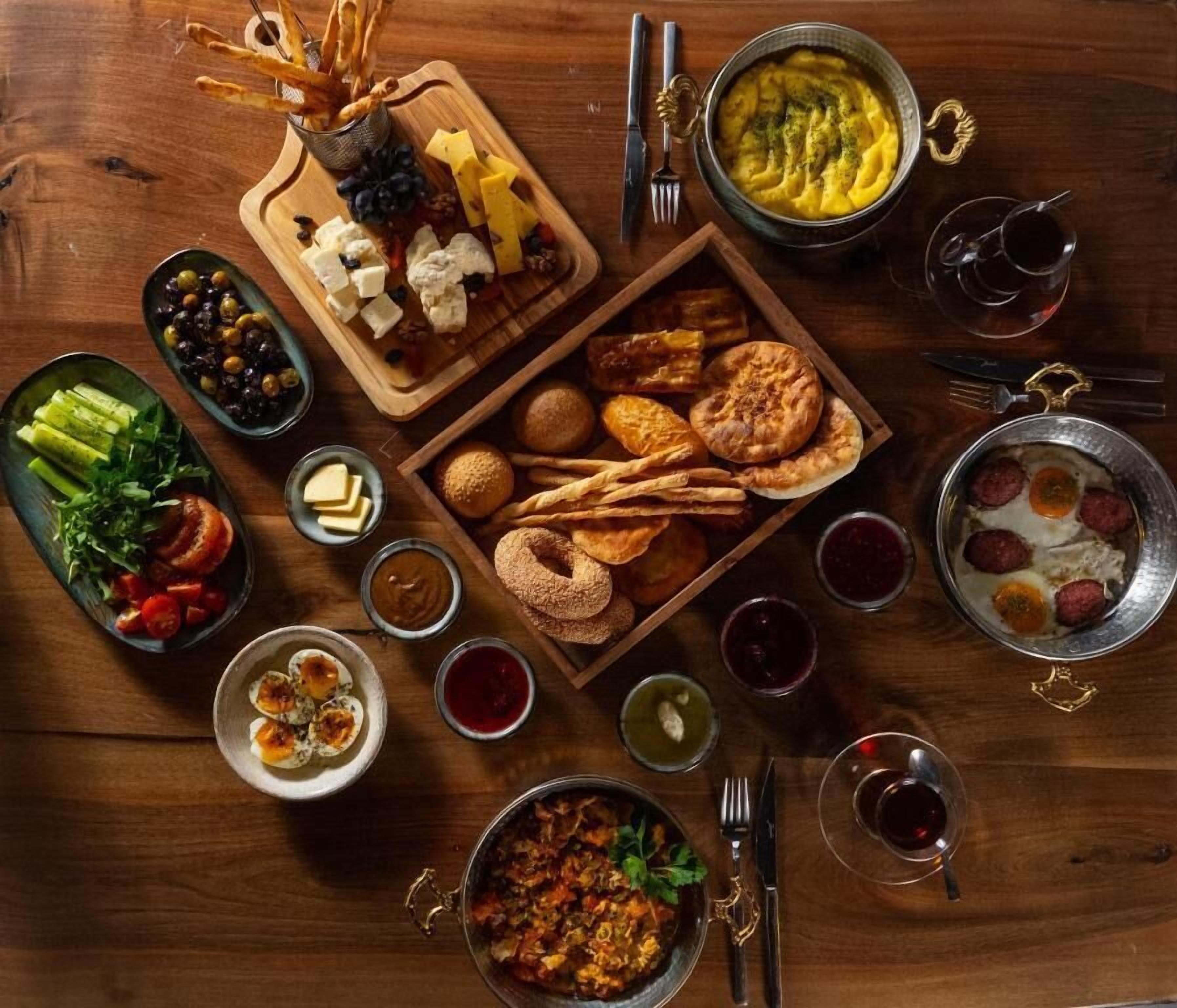 A variety of foods and drinks arranged on a wooden table, including cheeses, bread, olives, and a bowl of soup.