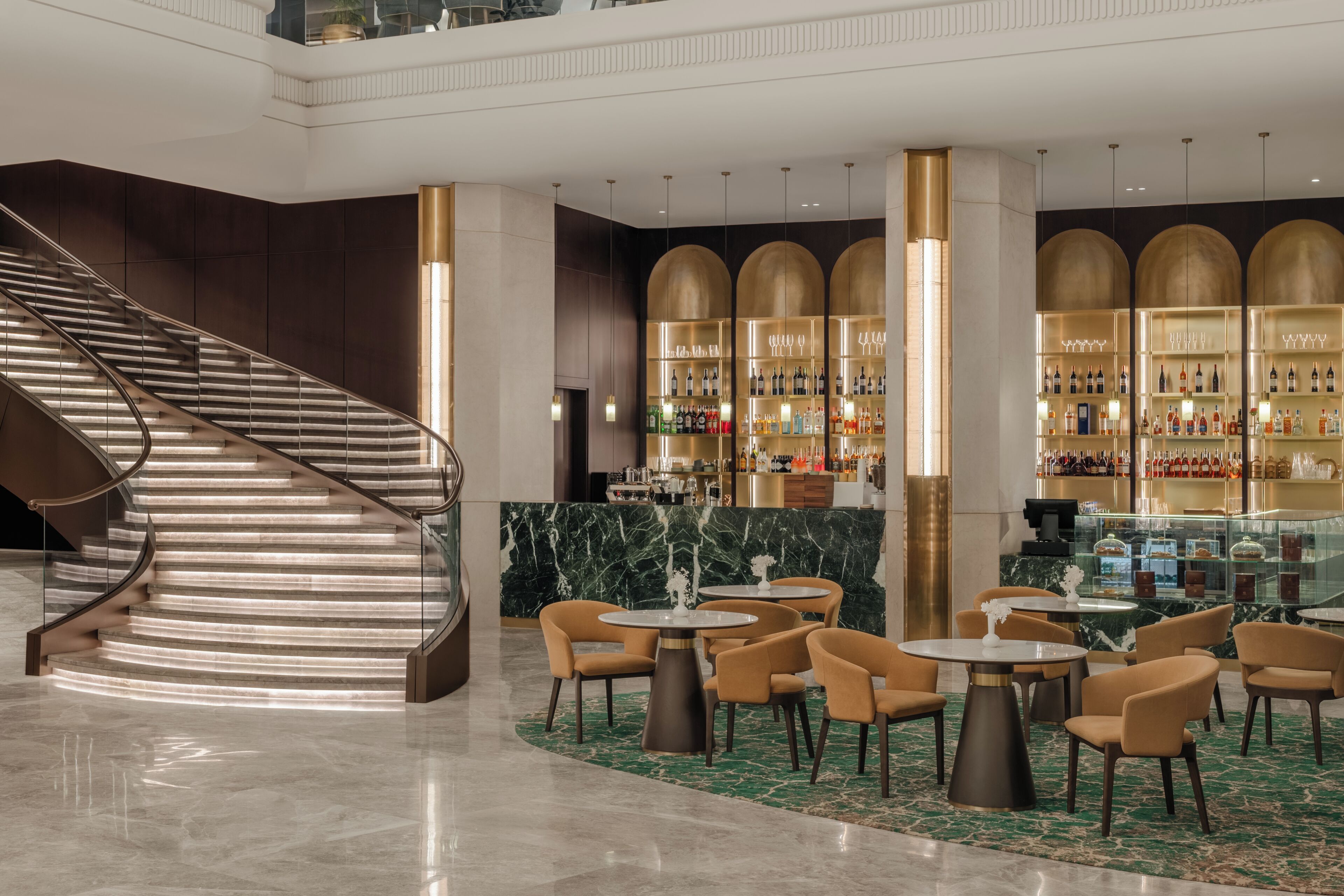 Un moderno vestíbulo de hotel con una escalera de mármol, una zona de bar con estantes llenos de botellas y varias mesas redondas con sillas.