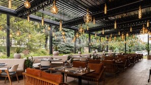 An indoor dining area with wooden tables and chairs, hanging pendant lights, and large windows overlooking a garden. - Hotel Kazakhstan (Almaty)