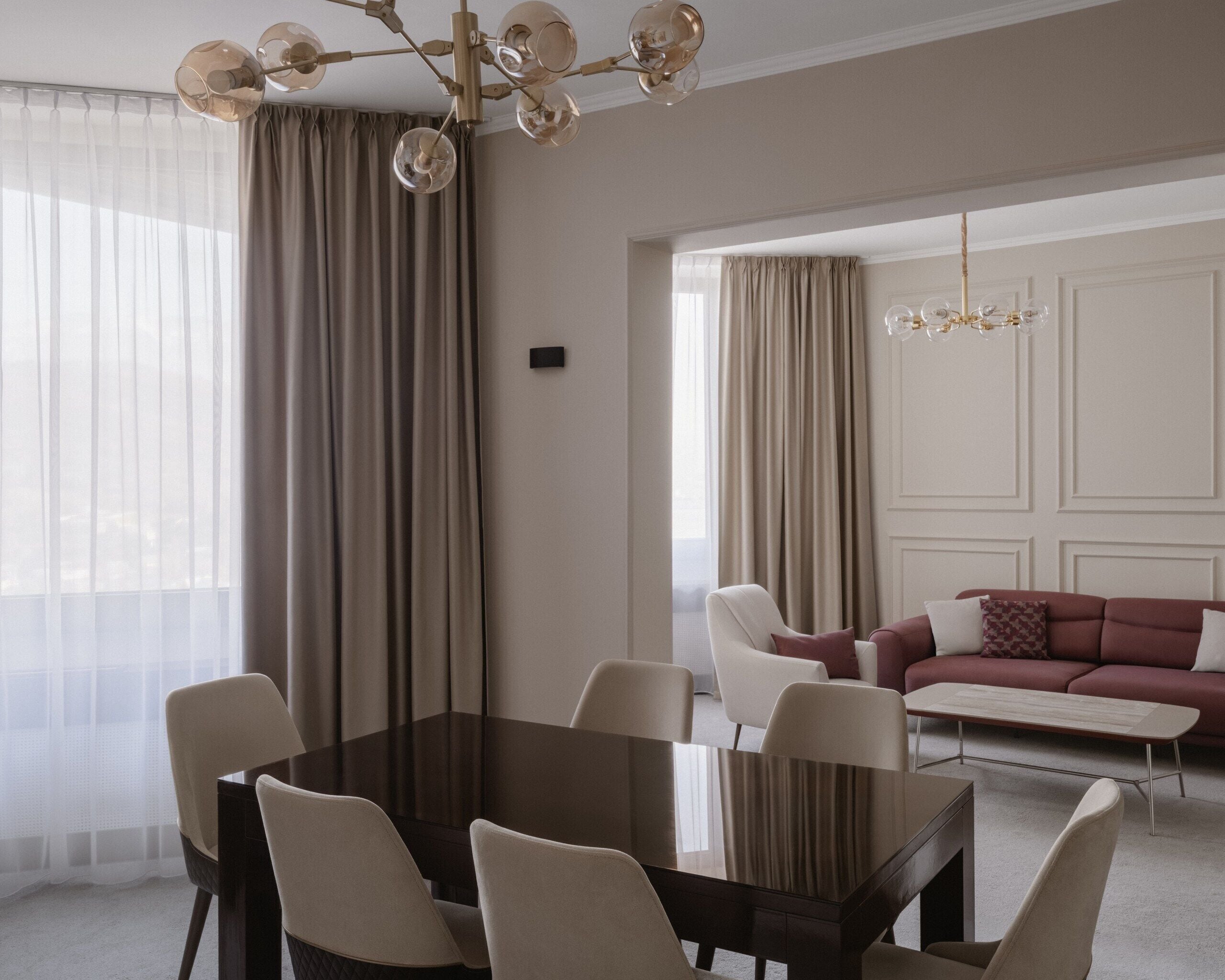 A modern living room with a dining area, featuring a dark wooden table, beige chairs, a red sofa, and a chandelier.