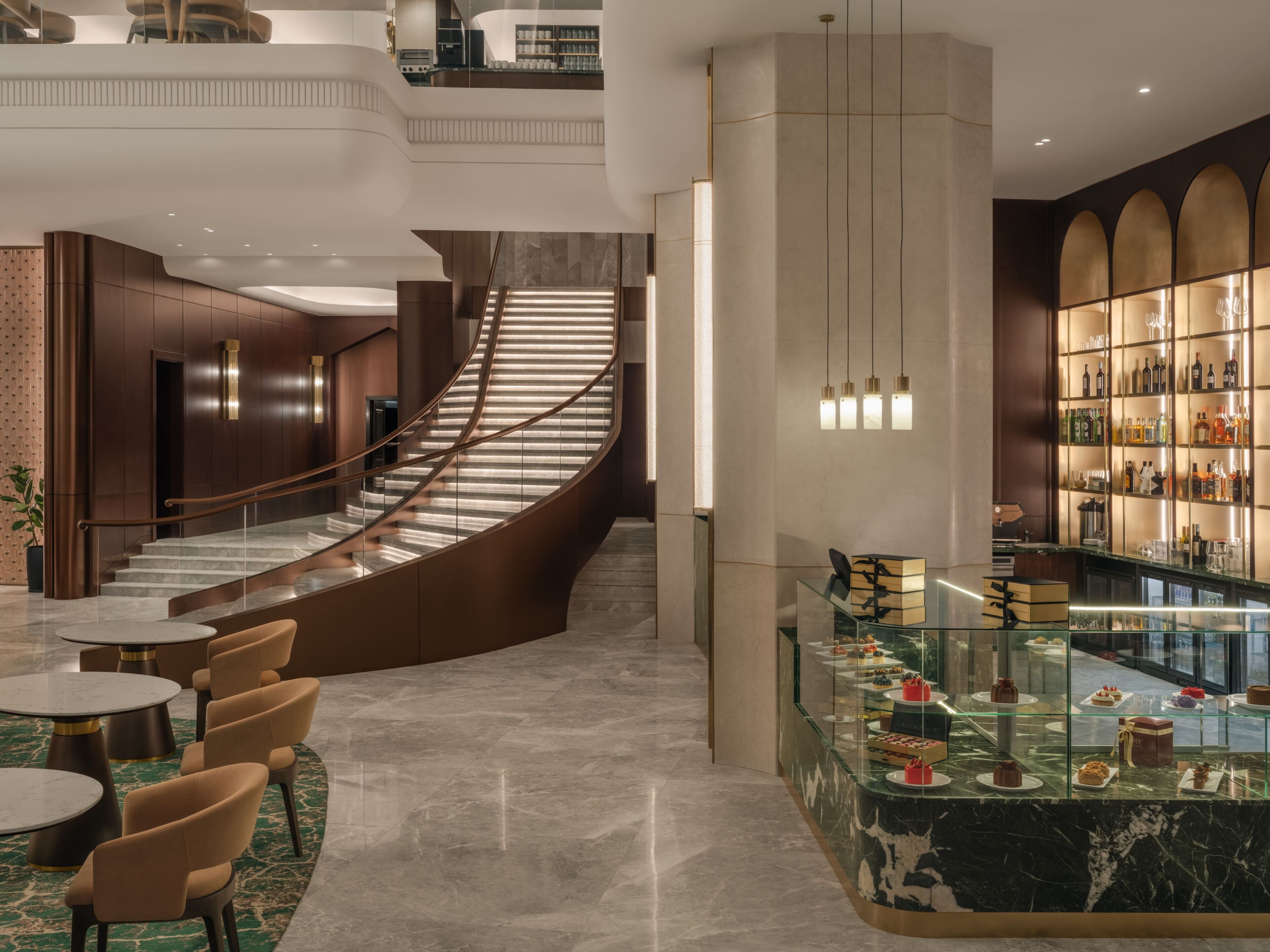 A modern hotel lobby with a curved staircase, a glass display case of desserts, and a bar area.
