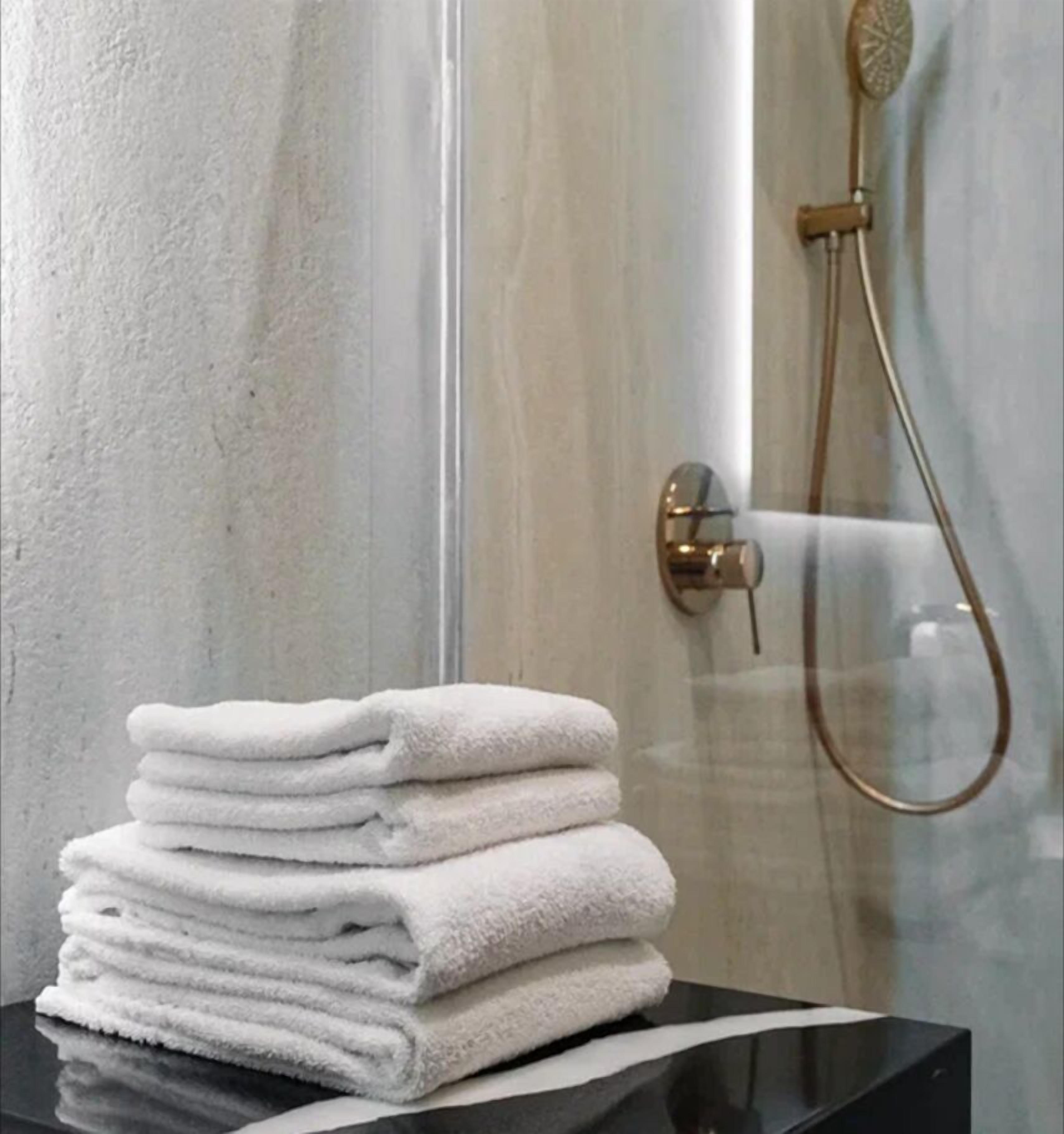 Una pila de toallas blancas sobre una superficie negra en un baño con mampara de vidrio y ducha con cabezal dorado.