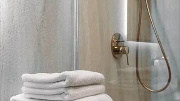 Una pila de toallas blancas sobre una superficie negra en un baño con mampara de vidrio y ducha con cabezal dorado.