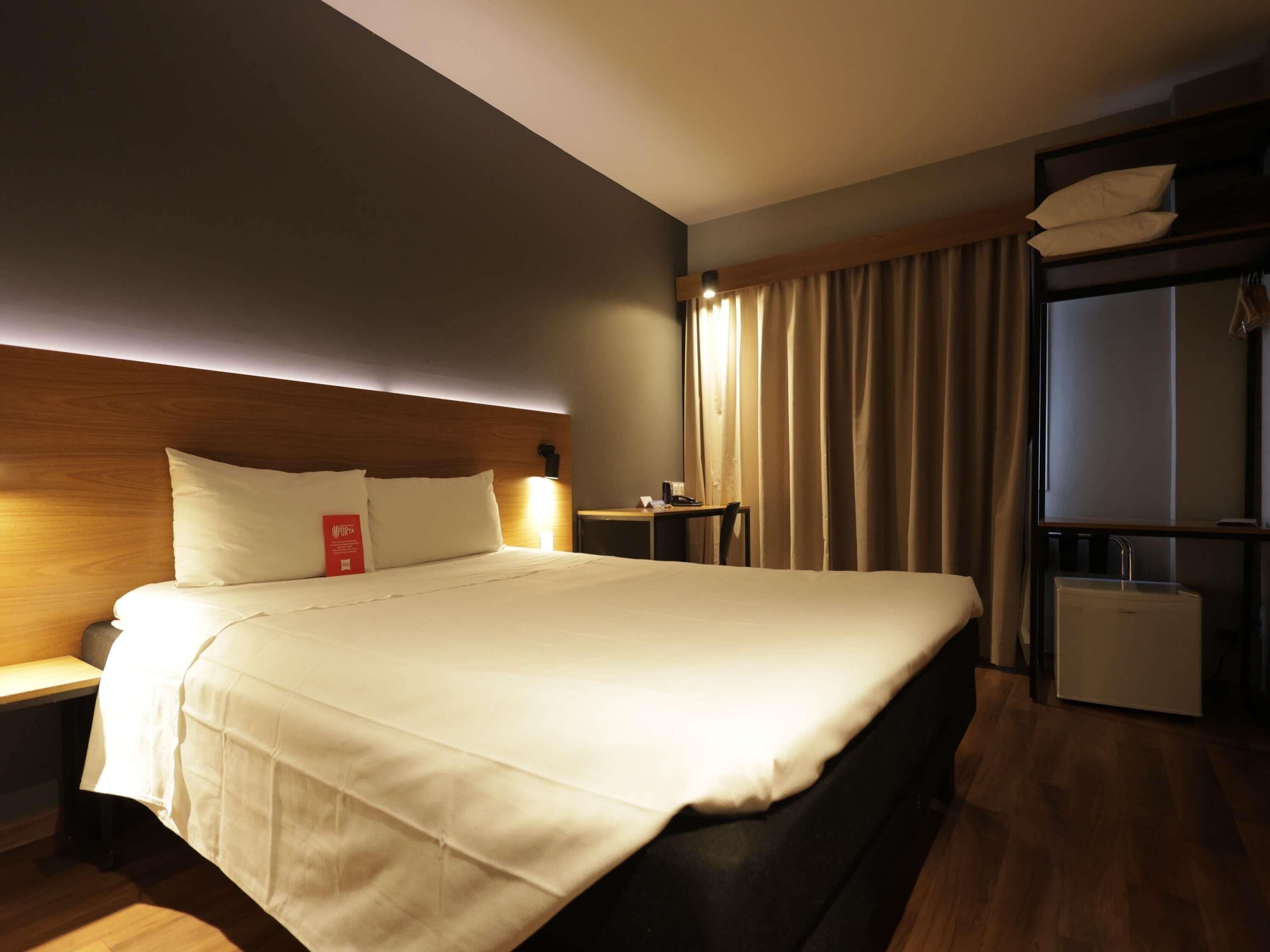 A hotel room with a large bed, bedside tables, a desk, a chair, a wardrobe, and a window with curtains.