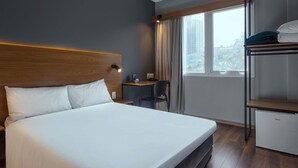 Une chambre d’hôtel équipée d’un lit, d’un bureau, d’une chaise, d’une armoire et d’une fenêtre donnant sur des immeubles.