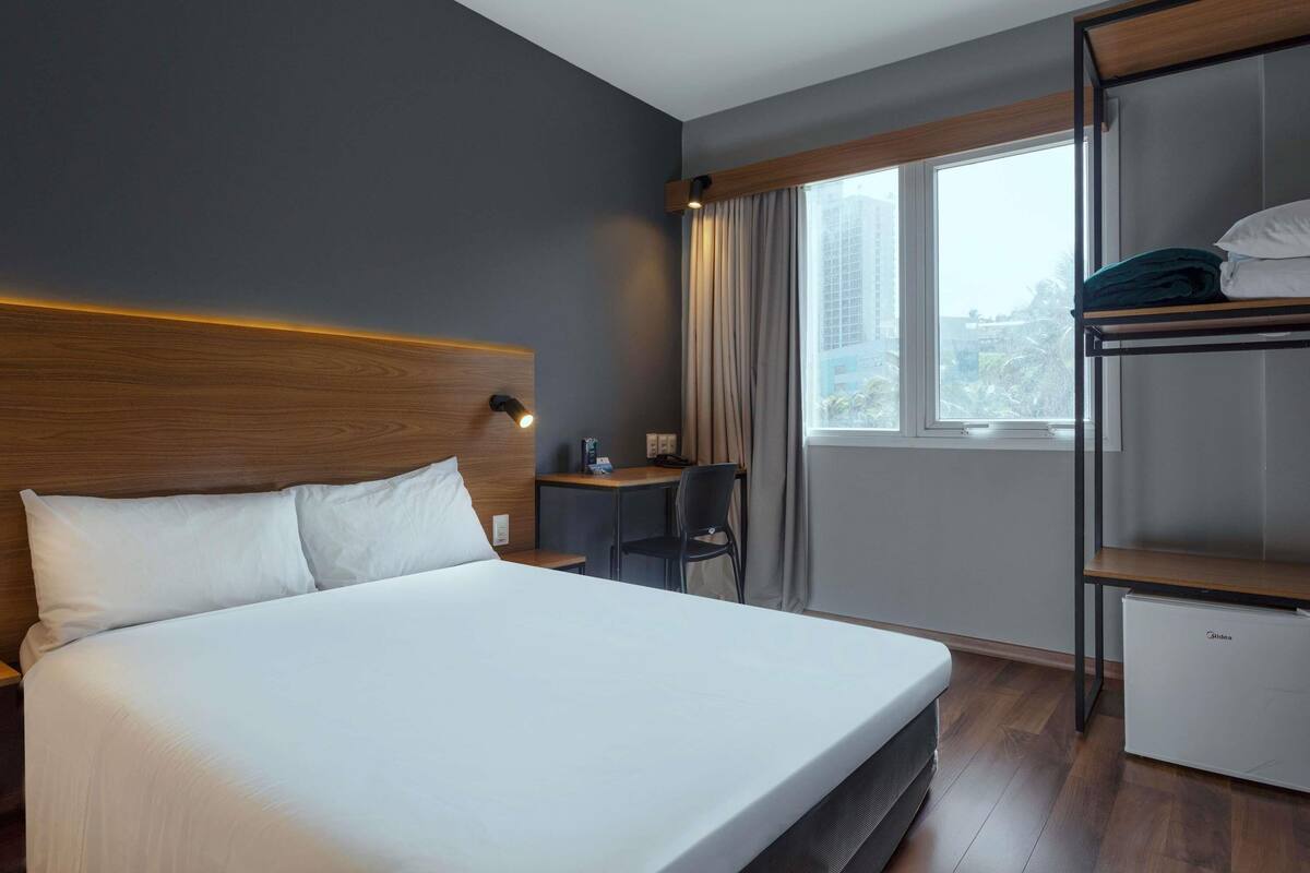 Une chambre d’hôtel équipée d’un lit, d’un bureau, d’une chaise, d’une armoire et d’une fenêtre donnant sur des immeubles.
