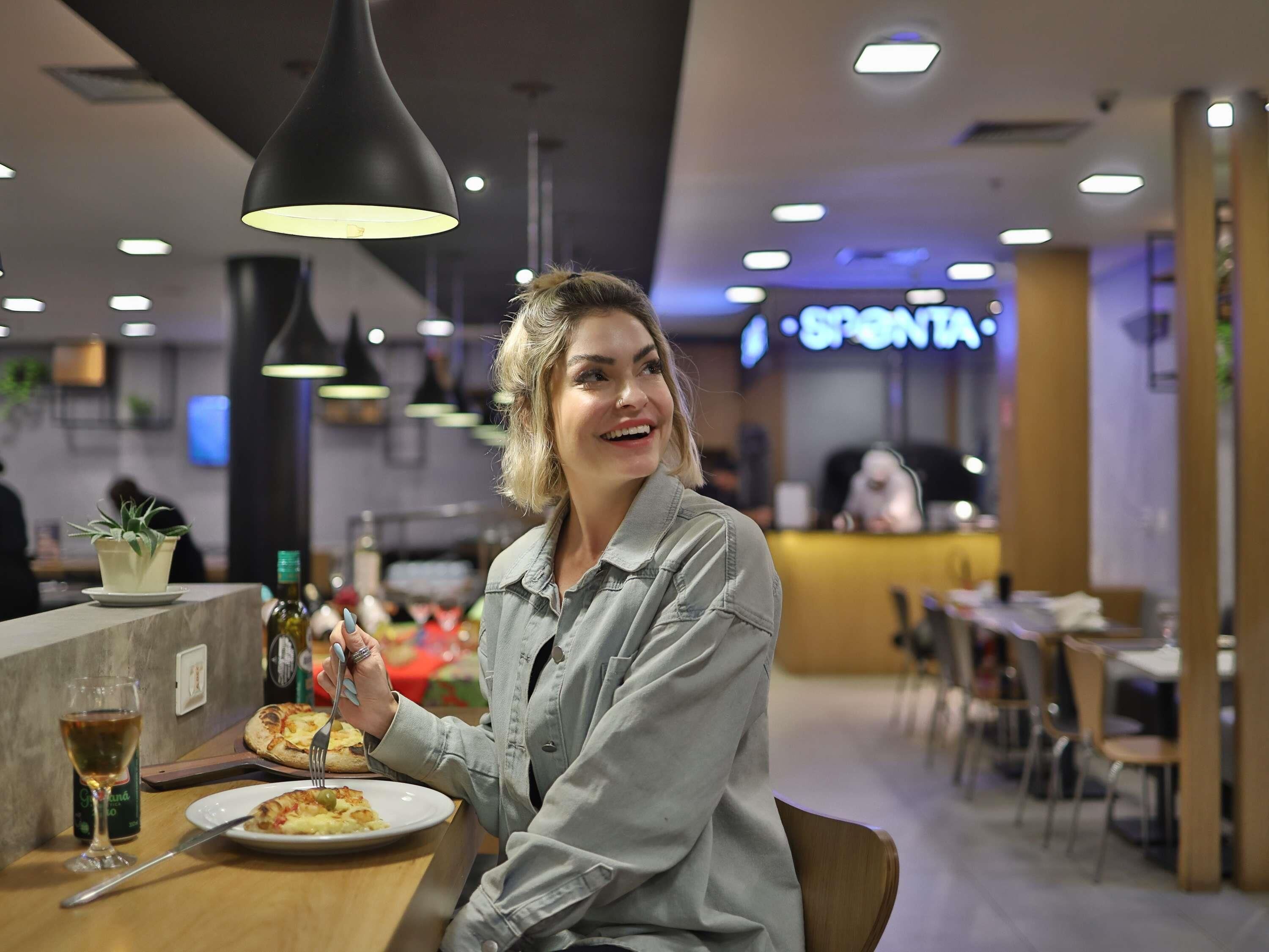 A person seated at a table with a plate of food, a glass of drink, and a bottle of olive oil. The setting appears to be a restaurant or cafe with a modern interior, including pendant lighting and a visible counter area.