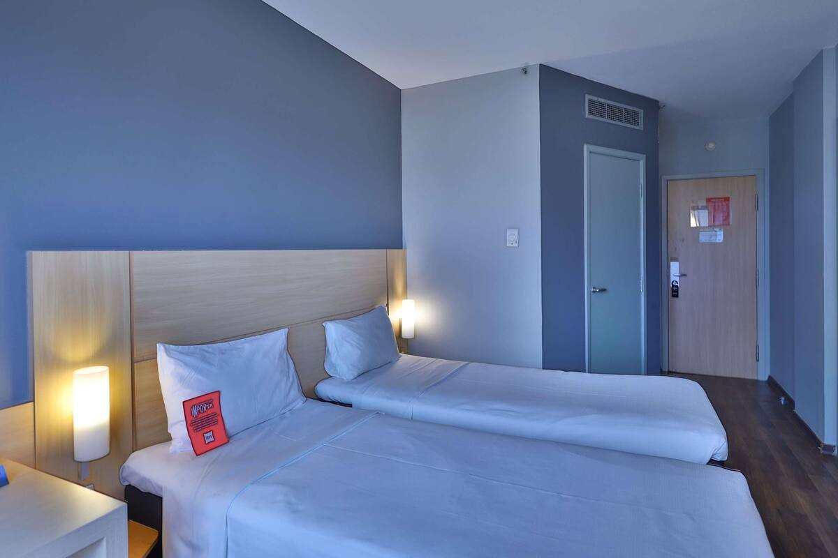 Une chambre d’hôtel avec deux lits, une table de chevet, une lampe fixée au mur, une porte avec un avis affiché et un plancher en bois.